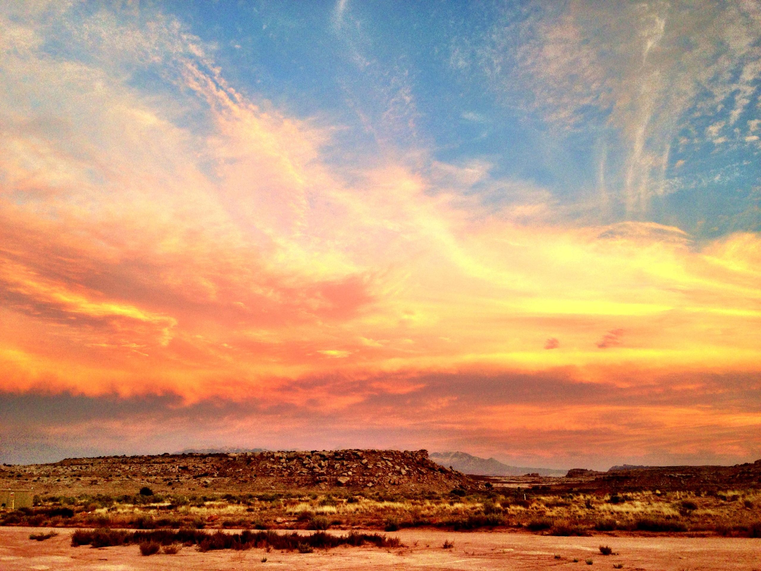 A vibrant sunset illuminates the sky with shades of orange, pink, and blue, casting a warm glow over a rugged landscape. In the foreground, sparse vegetation and rocky terrain are visible beneath a dramatic sky filled with wispy clouds. Sovereign Single Track mountain bike trail.