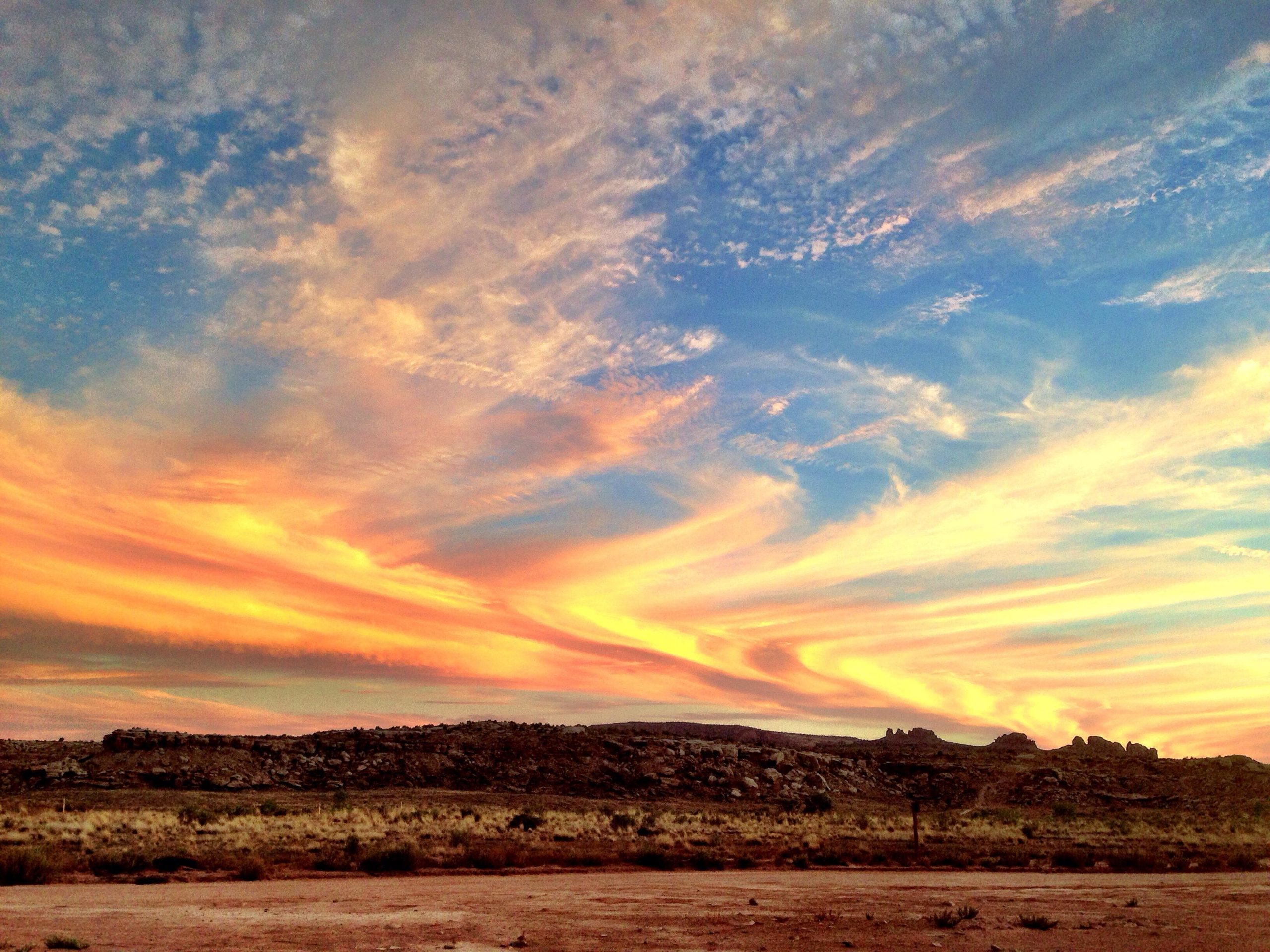 A vibrant sunset sky with swirling clouds in shades of orange, pink, and blue, over a rocky landscape. The foreground features sparse vegetation and dirt, while the horizon is marked by distant rock formations beneath a textured, colorful sky. Sovereign Single Track mountain bike trail.