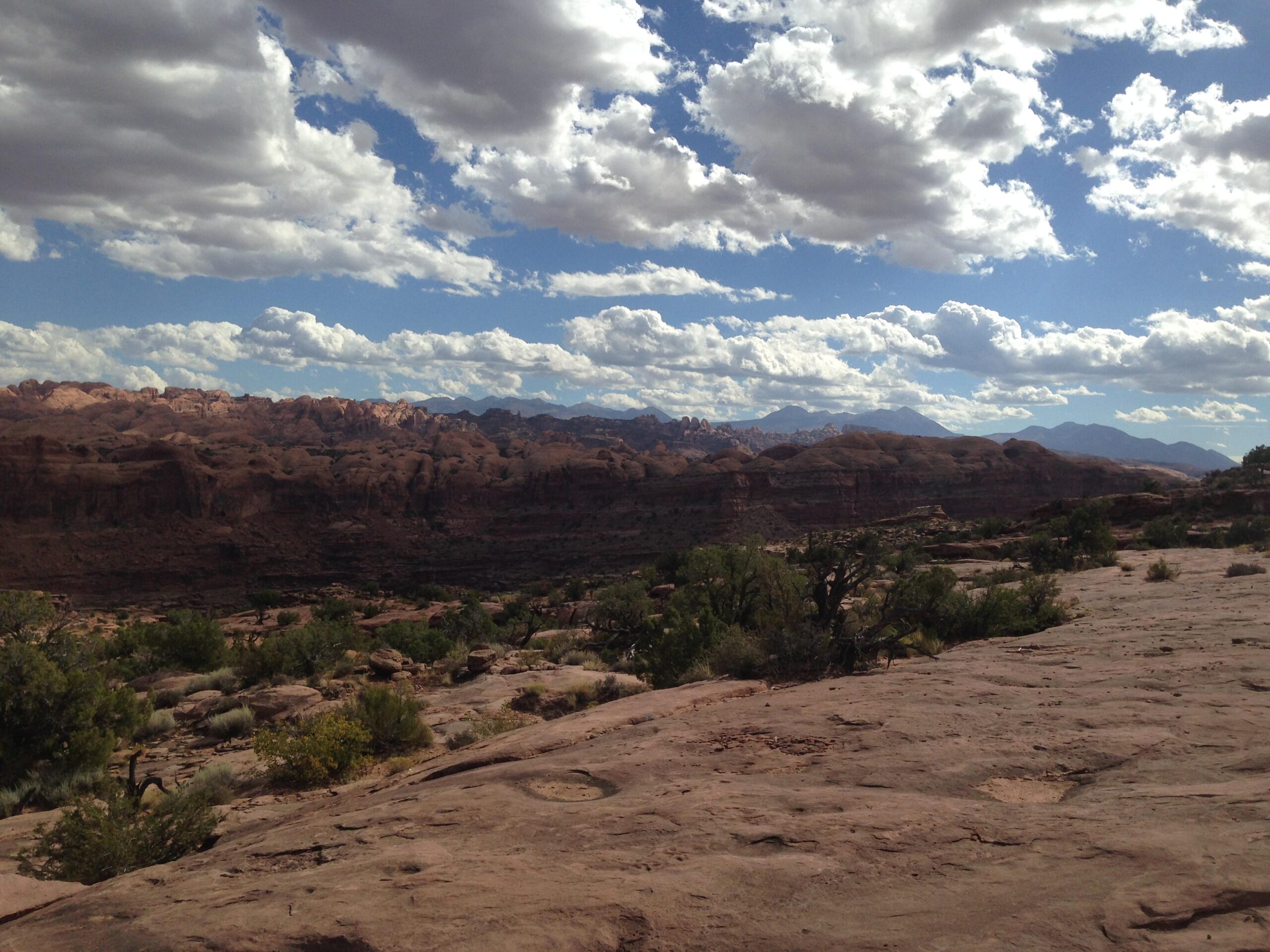 A panoramic view of a rocky desert landscape under a partly cloudy sky, featuring layered rock formations and scattered vegetation. The scene captures distant mountain ranges and a dramatic cloud formation overhead, illustrating the beauty of the natural environment. Hymasa mountain bike trail.