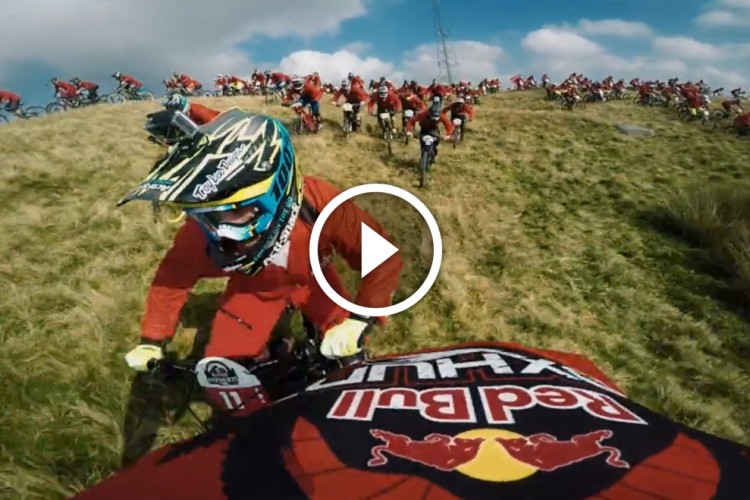 A mountain biker in a colorful helmet and gear rides downhill, with a group of riders in red apparel visible in the background on a grassy hill. The scene is dynamic and captures the excitement of a biking event, with the Red Bull logo prominently displayed on the bike.