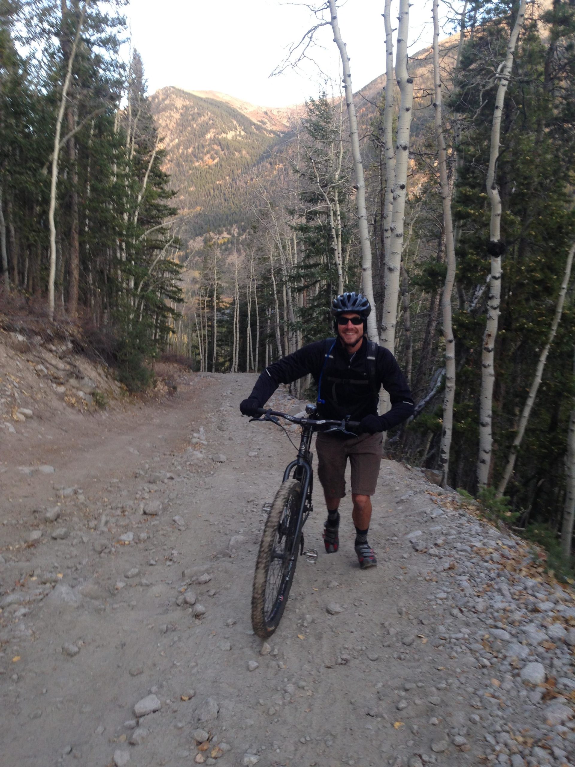 A mountain biker pushing their bike up a gravel trail surrounded by tall trees and mountains in the background. The scene captures a sense of adventure and the beauty of nature in an outdoor setting. Baldwin Creek Road / FS #277 mountain bike trail.