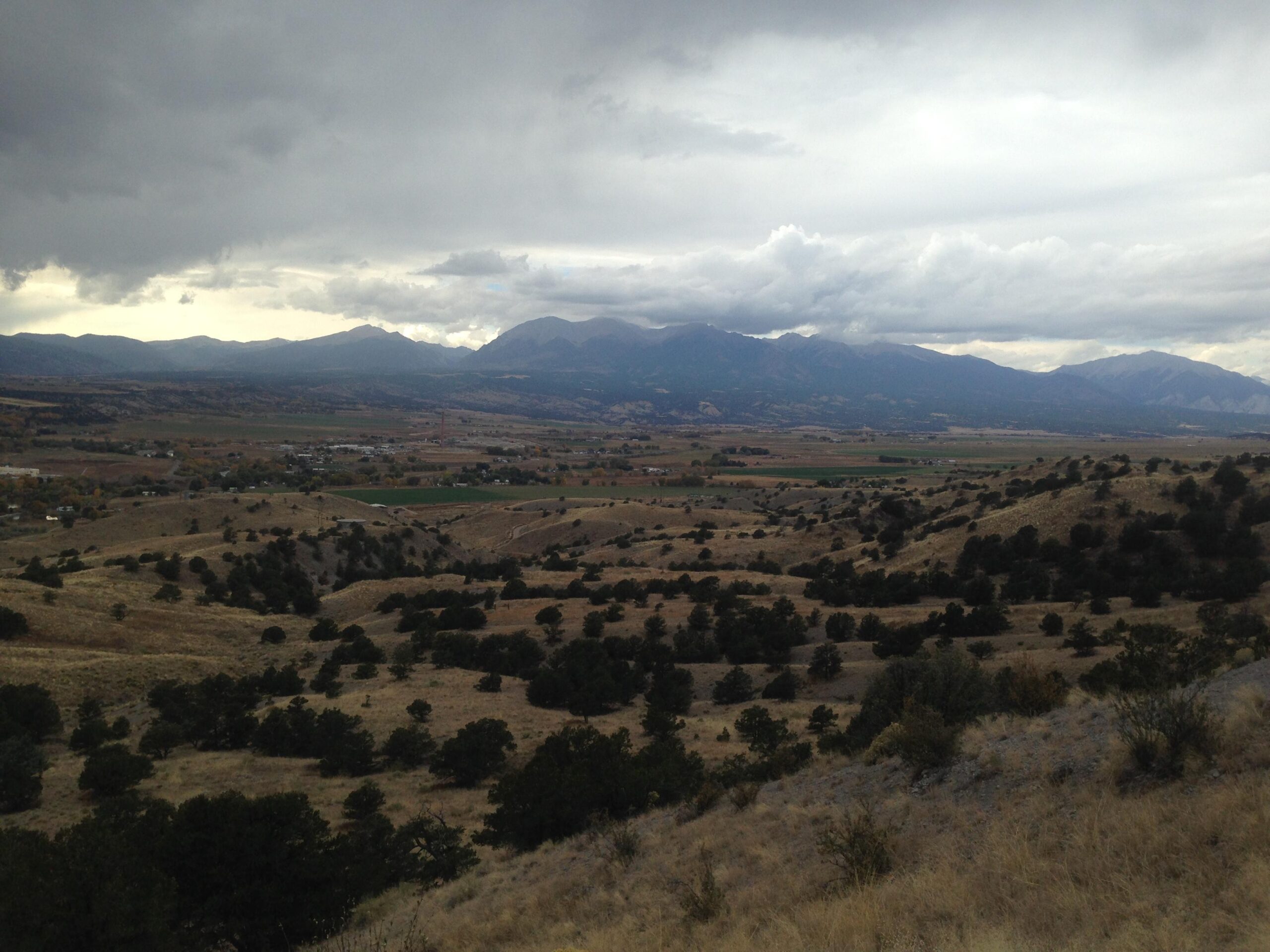 A panoramic view of a mountainous landscape under a cloudy sky. Rolling hills covered in dry grass and scattered trees extend into the distance, leading to a valley with patches of green fields and a small town. The mountains in the background are partially shrouded in clouds, creating a dramatic and serene atmosphere. Arkansas Hills mountain bike trail.