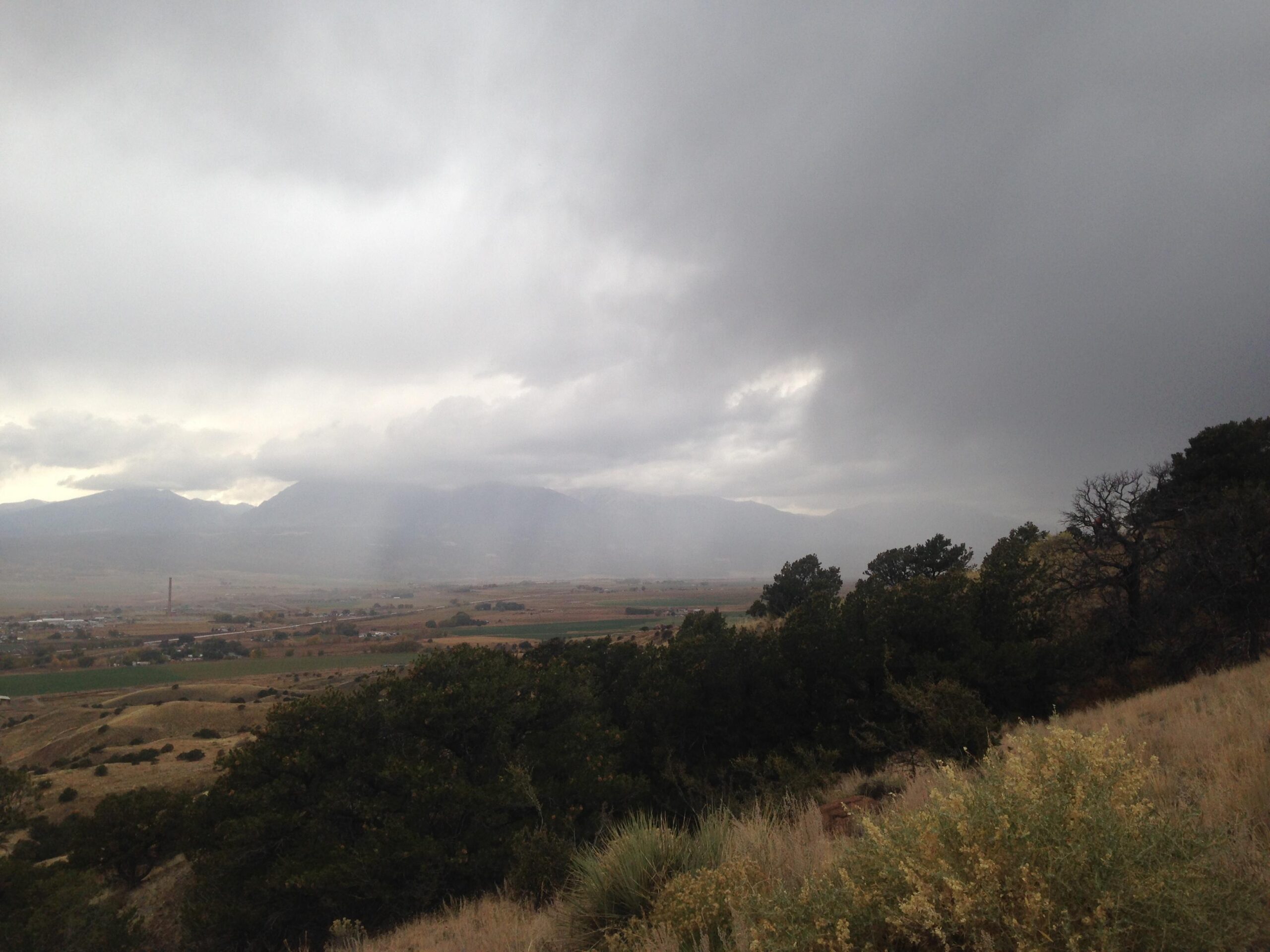 A cloudy landscape view featuring dark gray skies, distant mountains shrouded in mist, and green fields below. The foreground includes shrubs and dry grass, with a glimpse of a small town visible in the valley. The atmosphere suggests an impending rain. Arkansas Hills mountain bike trail.