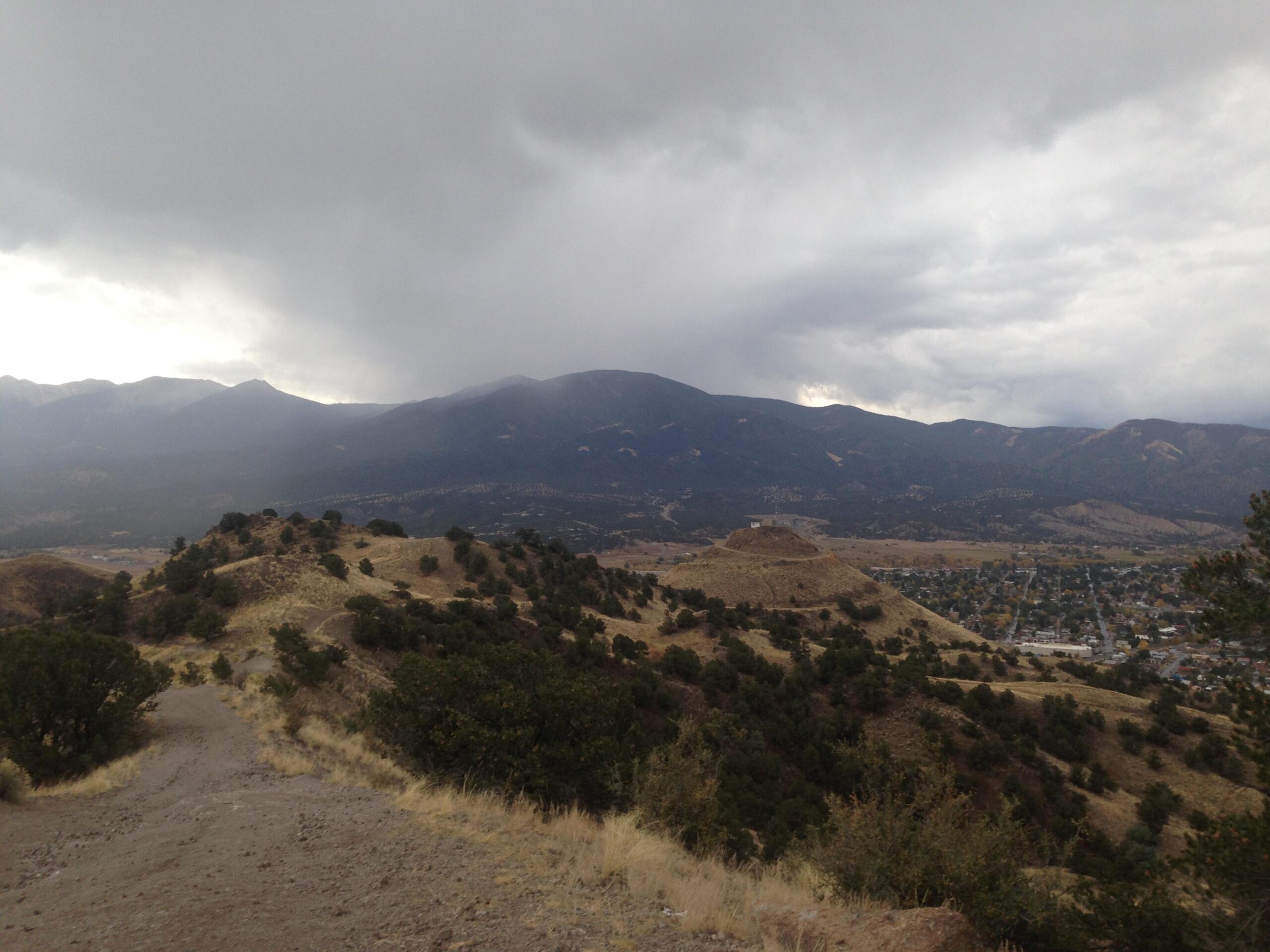 A scenic view of rolling hills and mountains under a cloudy sky, with a small town visible in the valley below. The landscape features dry grasses and scattered trees, creating a rugged and natural atmosphere. Arkansas Hills mountain bike trail.