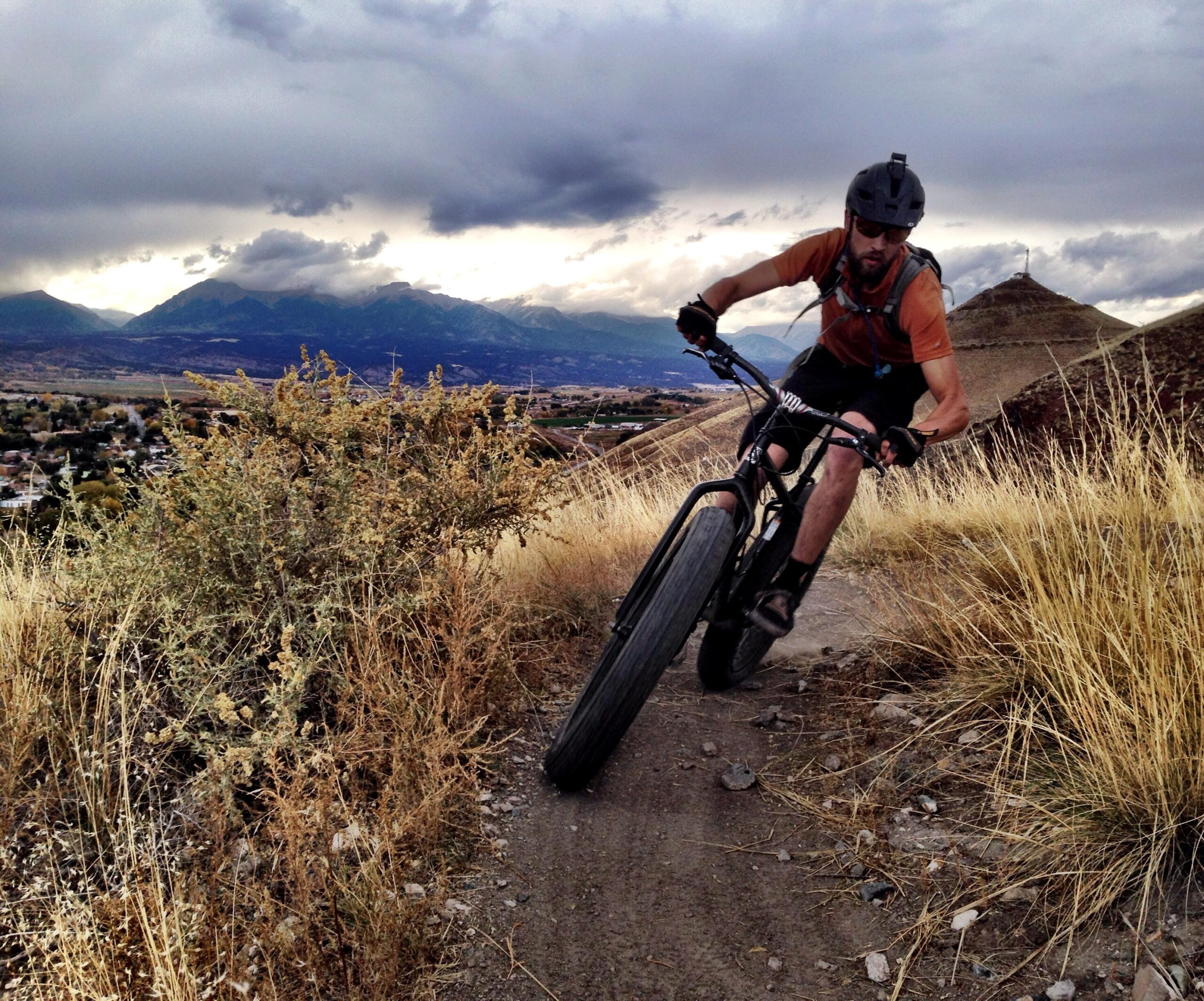 A mountain biker navigating a dirt trail on a hillside, surrounded by dry grass and shrubs, with a mountainous landscape and stormy clouds in the background. The biker is leaning into a turn, wearing a helmet and protective gear. Arkansas Hills mountain bike trail.