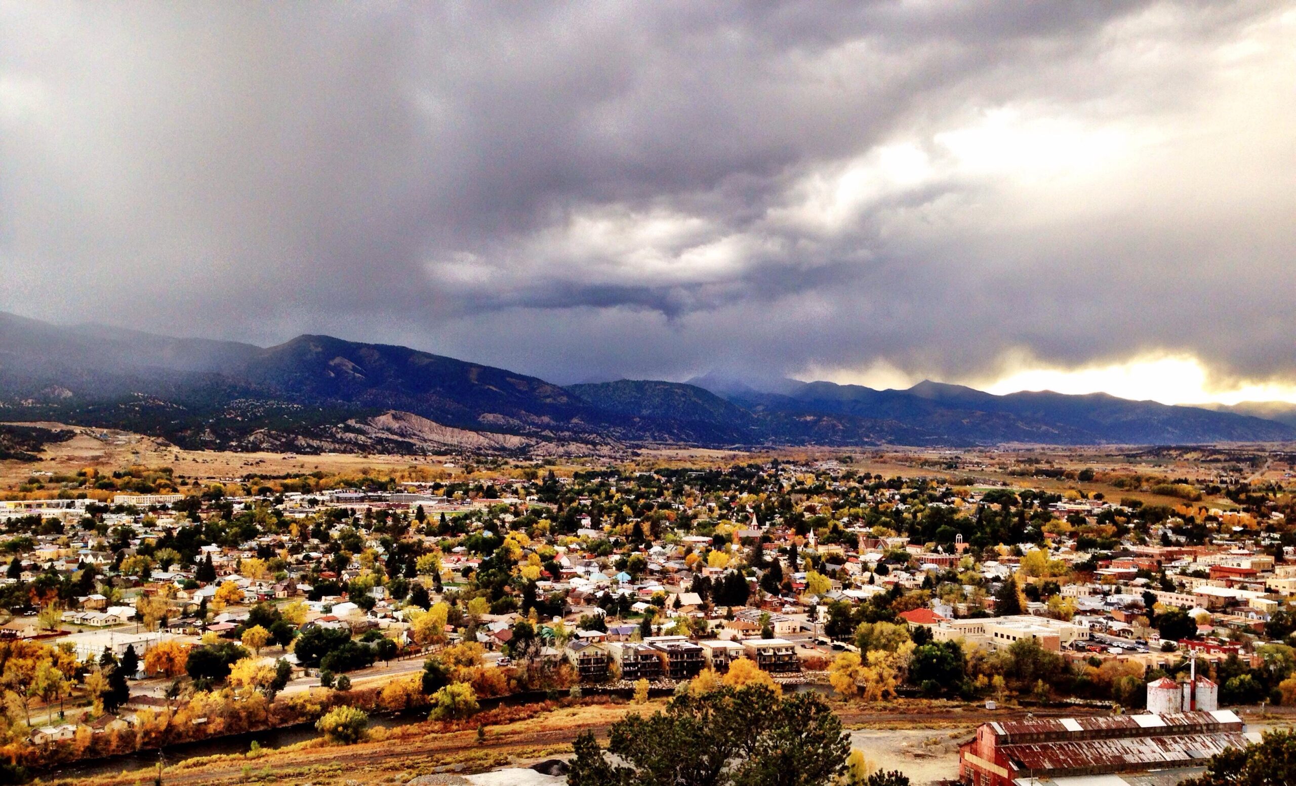 A panoramic view of a small town nestled in a valley, surrounded by mountains under a cloudy sky. The landscape features a mix of residential buildings and trees, with autumn foliage adding vibrant colors. The foreground includes a structure with a rusted roof, while the background showcases rolling hills and dark storm clouds threatening rain. Arkansas Hills mountain bike trail.