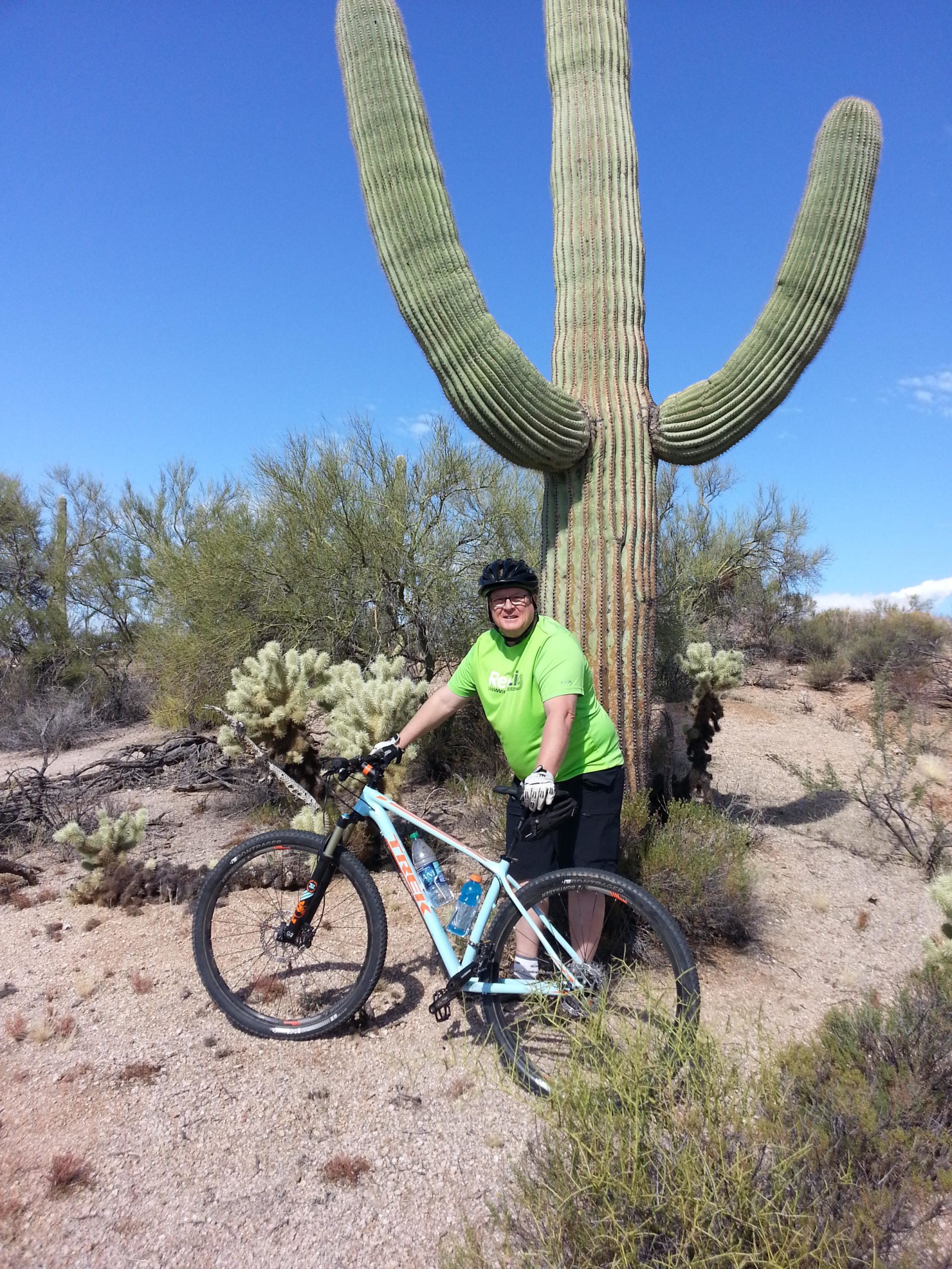 A person wearing a bright green cycling shirt and a black helmet stands next to a large saguaro cactus while holding a mountain bike. The background features a desert landscape with sparse vegetation and a clear blue sky. Brown's Ranch to Granite Mountain mountain bike trail.