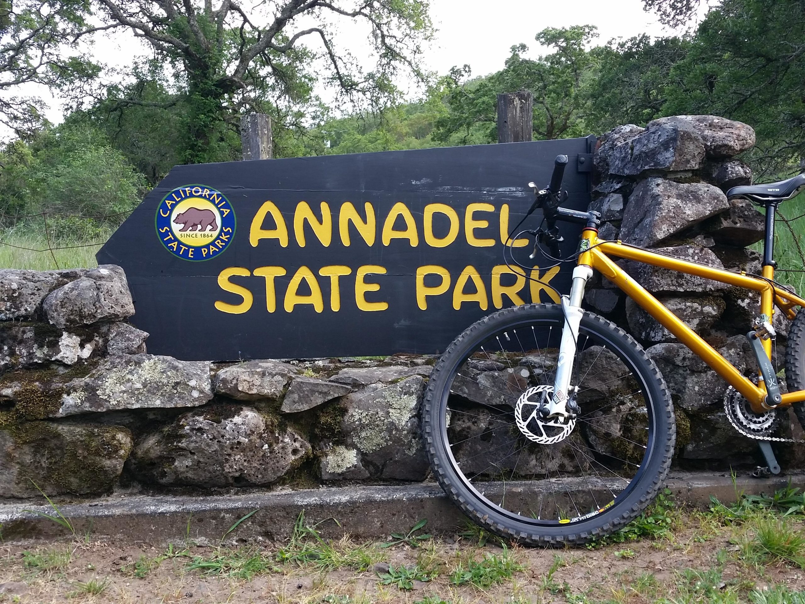 A mountain bike leaning against a large, colorful sign for Annadel State Park in California, surrounded by rocky landscaping and green trees. The sign features the park's name prominently in yellow letters, along with the California State Parks logo. Annadel State Park mountain bike trail.