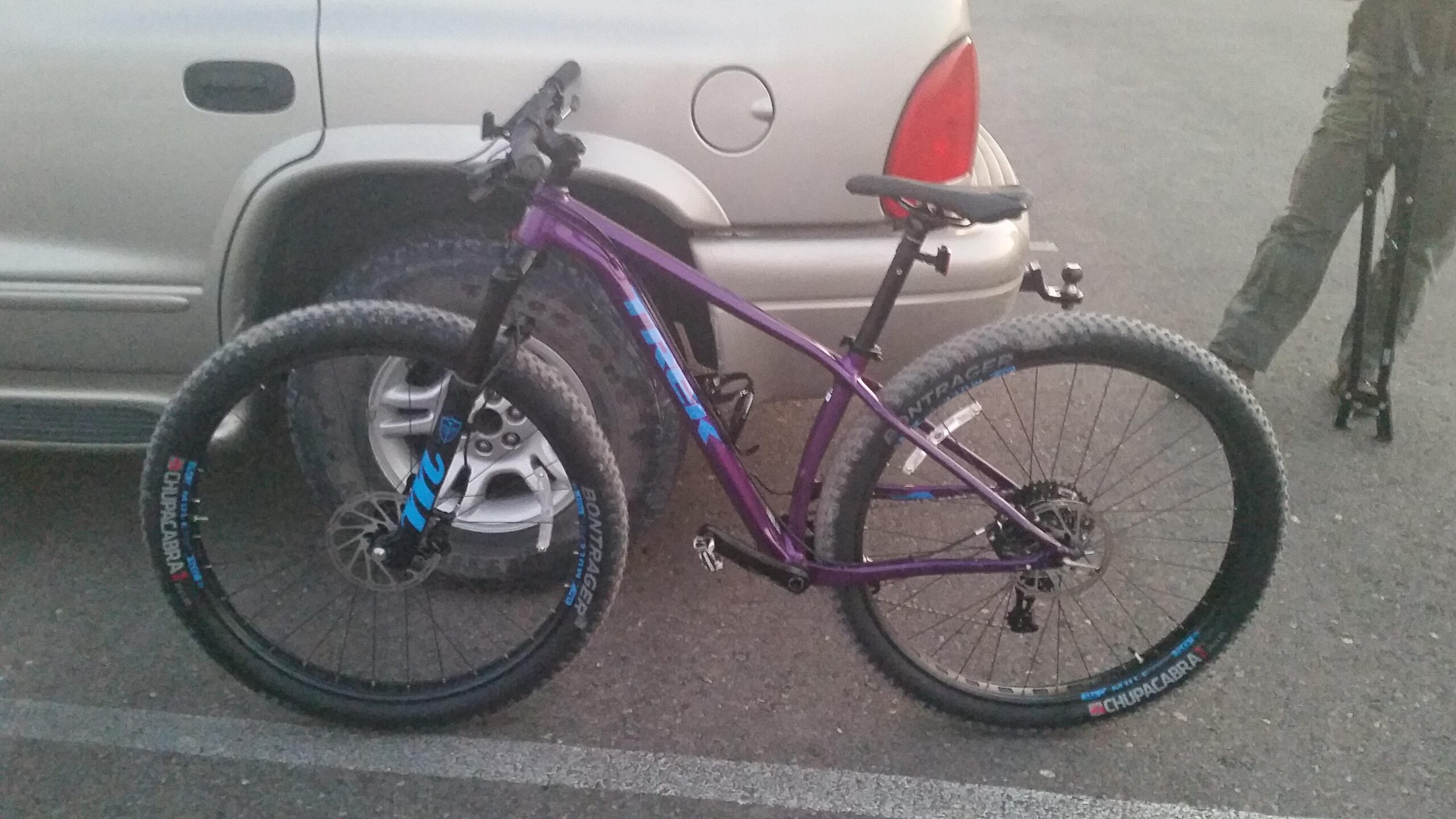 Trek Stache: A purple Trek mountain bike with thick tires parked next to a silver vehicle. The bike is positioned in a parking lot, showing its frame, wheels, and handlebars. In the background, part of a vehicle's wheel can be seen, and a person is standing nearby holding a bike pump.