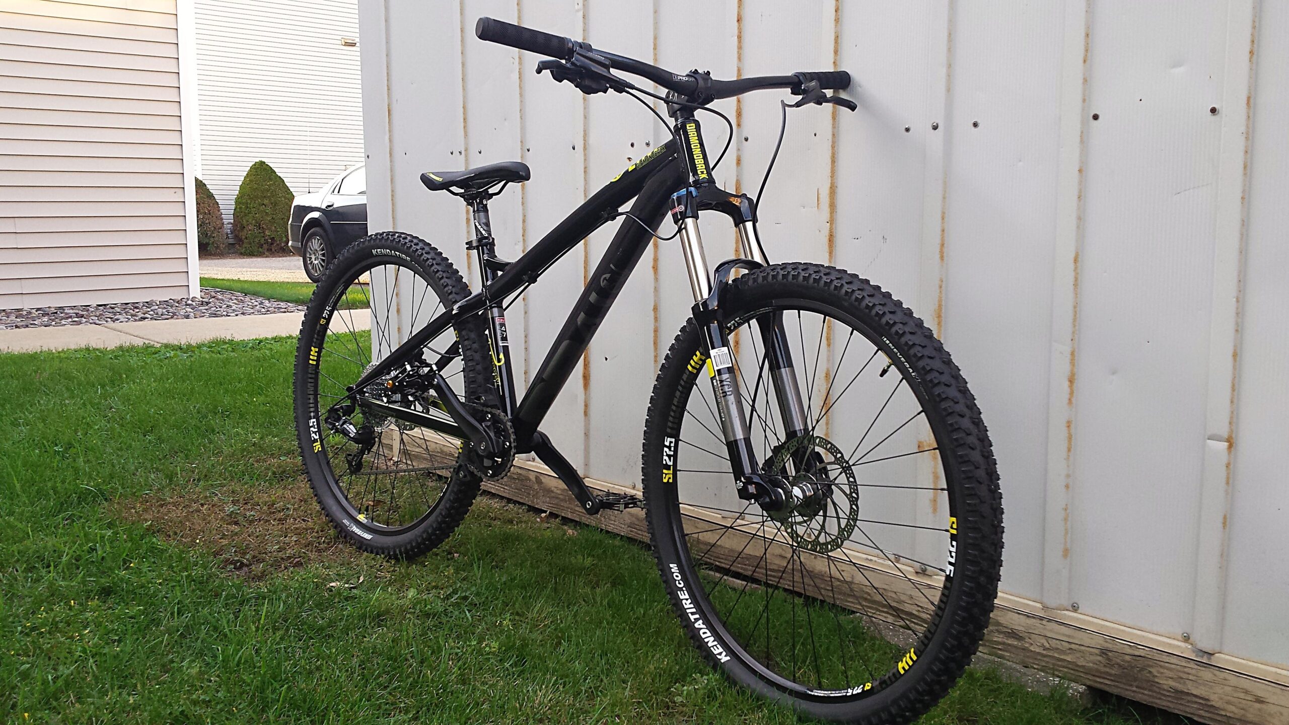 Diamondback Line: A black mountain bike with wide tires is positioned on grass next to a light-colored wall. The bike features a sturdy frame and front suspension, with visible gears and brakes. In the background, a portion of a car and some well-trimmed bushes can be seen.