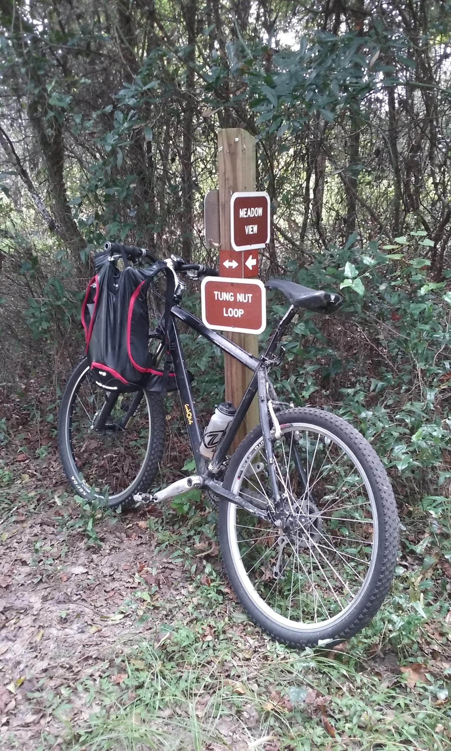 A mountain bike leaning against a trail sign directing to "Meadow View" and "Tung Nut Loop," surrounded by greenery and underbrush. San Felasco Hammock Preserve mountain bike trail.
