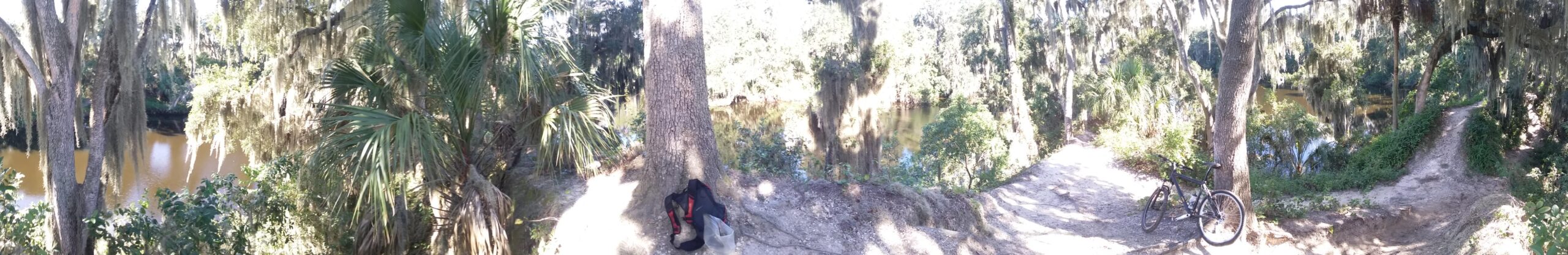A panoramic view of a serene riverside scene, featuring lush greenery, hanging moss, and a calm brown river. On the left, a black bicycle rests against a tree, while a backpack sits nearby on the ground. The sunlight filters through the leaves, creating a tranquil atmosphere. Loyce E. Harpe Park mountain bike trail.
