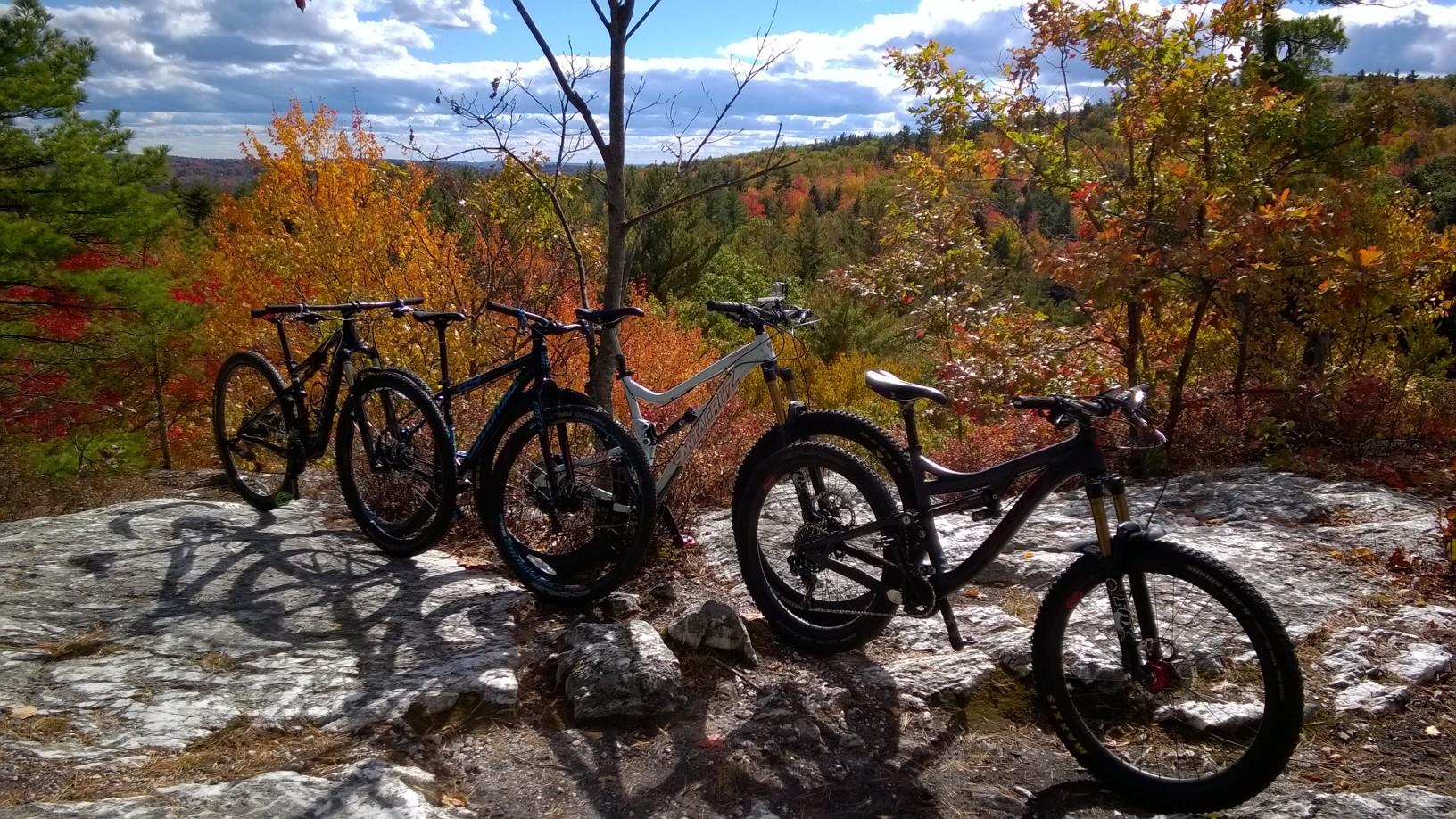 Four mountain bikes are parked on a rocky outcrop with a backdrop of colorful autumn foliage and a blue sky dotted with clouds. The bikes vary in design and color, showcasing their sturdy frames and features, while the vibrant leaves in shades of orange, red, and yellow create a picturesque outdoor setting. Bear Brook mountain bike trail.