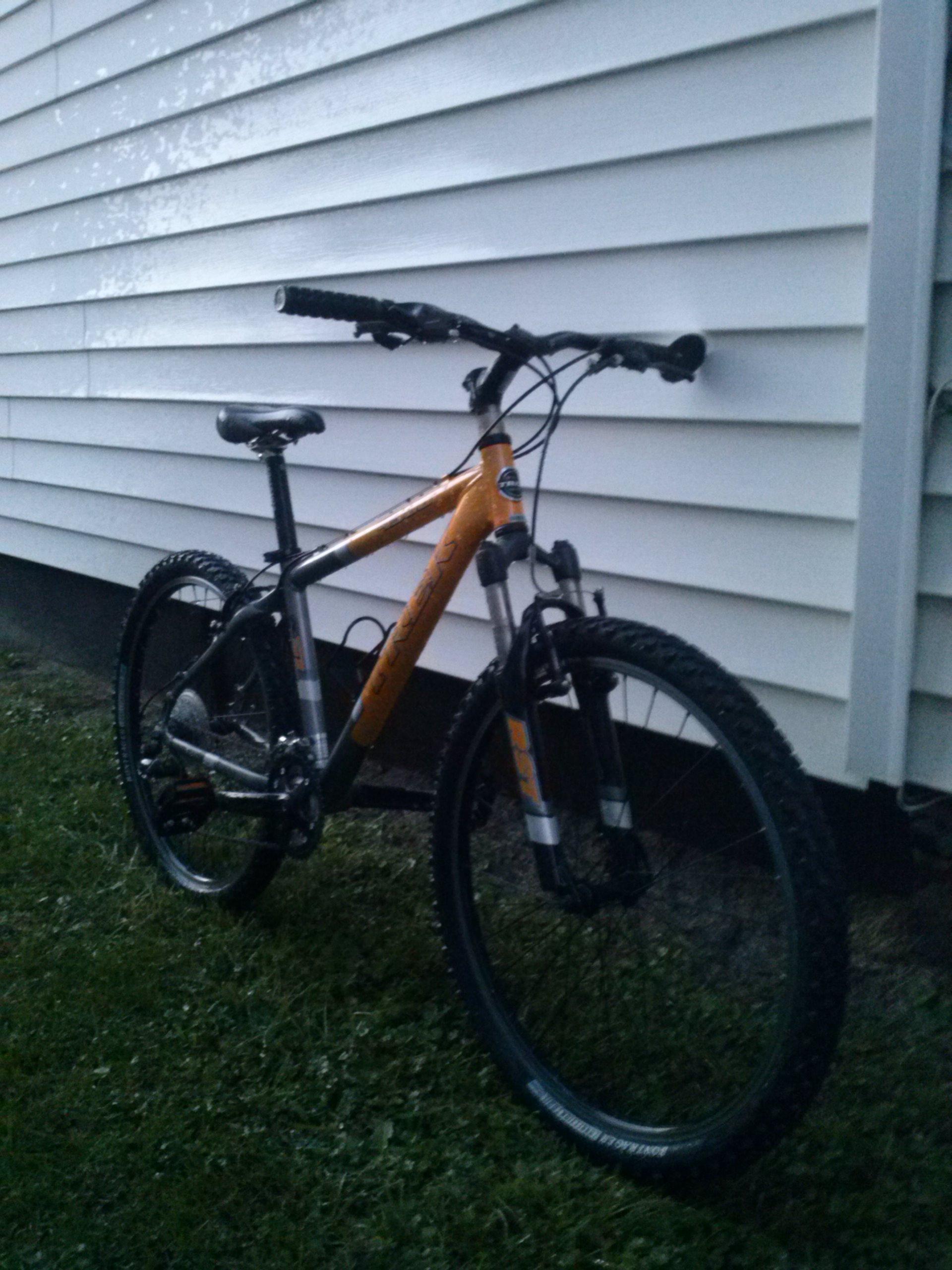 Trek 4300: Mountain bike leaning against a white, horizontally slatted wall with grass in the foreground. The bike has an orange and black frame, and features knobby tires designed for off-road use.