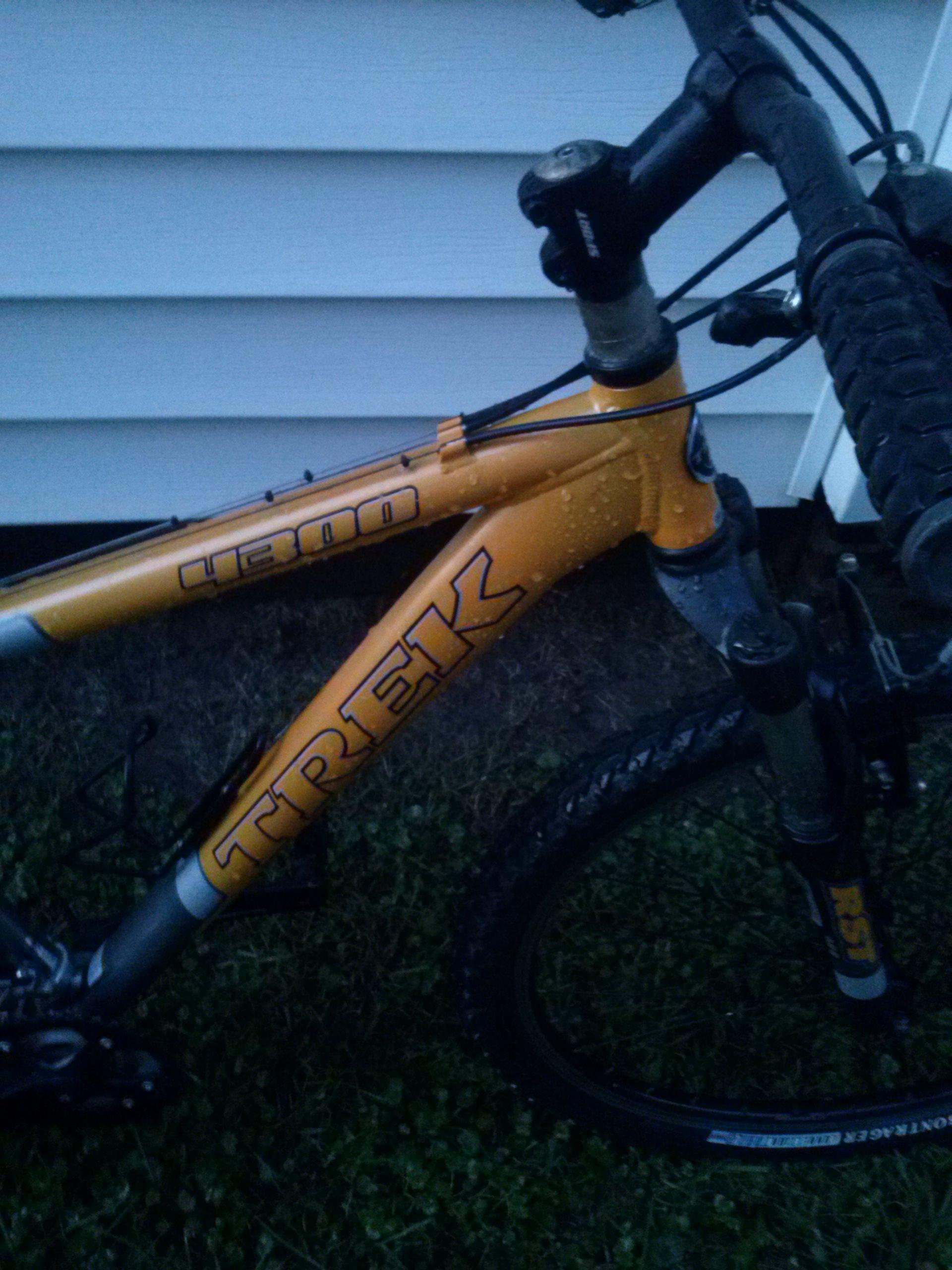 Trek 4300: A close-up view of a Trek mountain bike frame in a bright orange color, displaying the model number "4300." The bike is resting on grass, and the frame appears wet, suggesting recent exposure to rain. A portion of the handlebars and front tire is visible, set against a backdrop of a white wooden surface.
