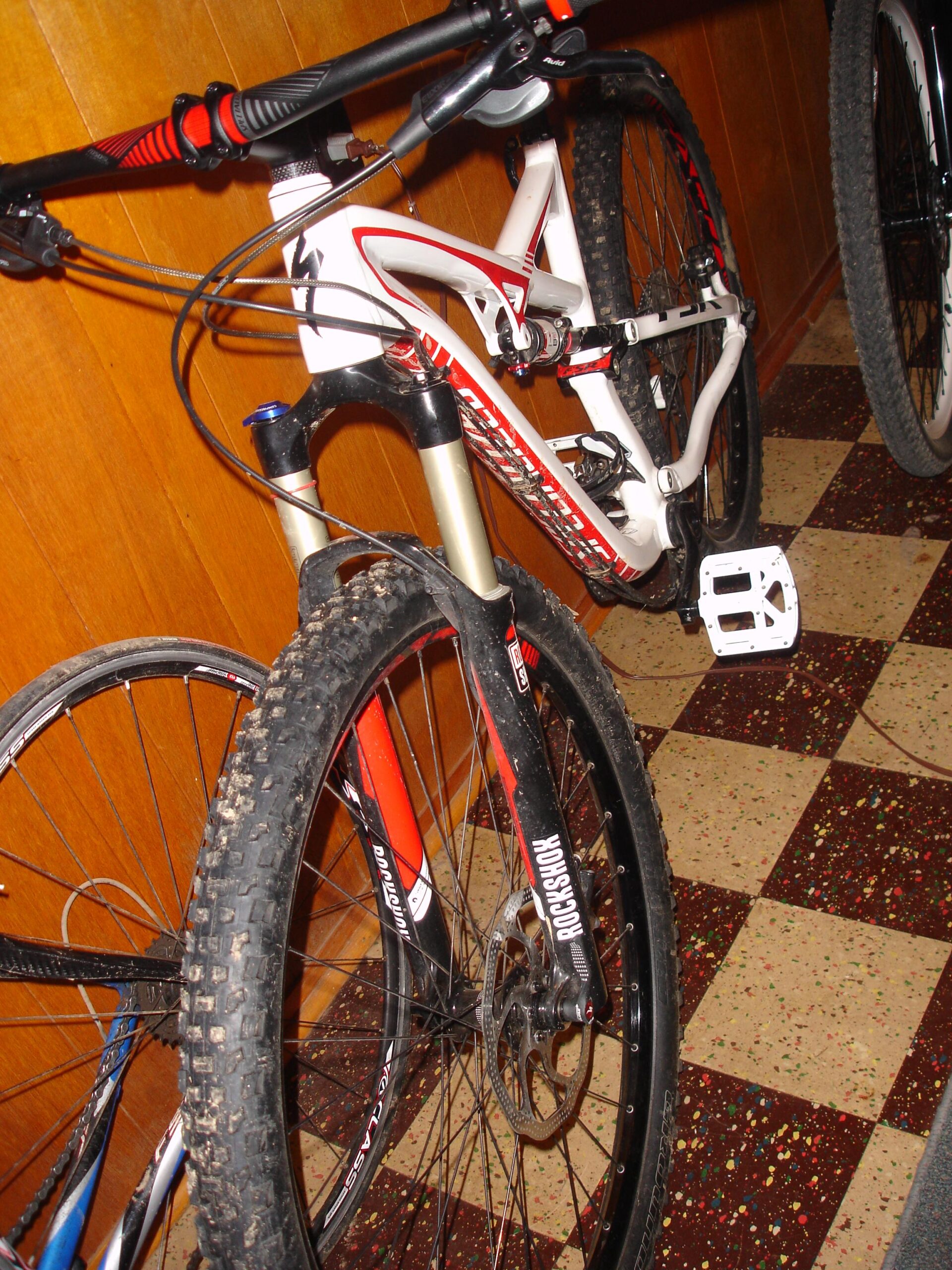 Specialized Camber Comp 29: A close-up image of a mountain bike, showcasing the front fork with suspension, handlebars, and a portion of the frame. The bike is positioned in a room with patterned tile flooring and wooden paneling in the background. The bike features a red and white color scheme, with visible dirt on the tires, indicating recent use.
