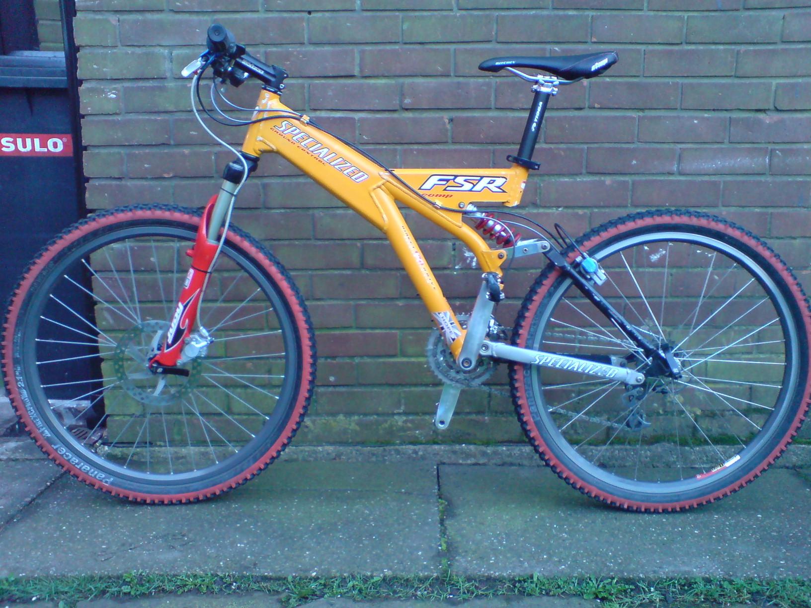 Specialized FSR: A bright orange and silver Specialized FSR mountain bike leaning against a brick wall. The bike features distinctive red accents on the front fork and tires, with a black saddle and handlebars. The ground is paved with square tiles, and there is a patch of grass visible in the lower corner of the image.