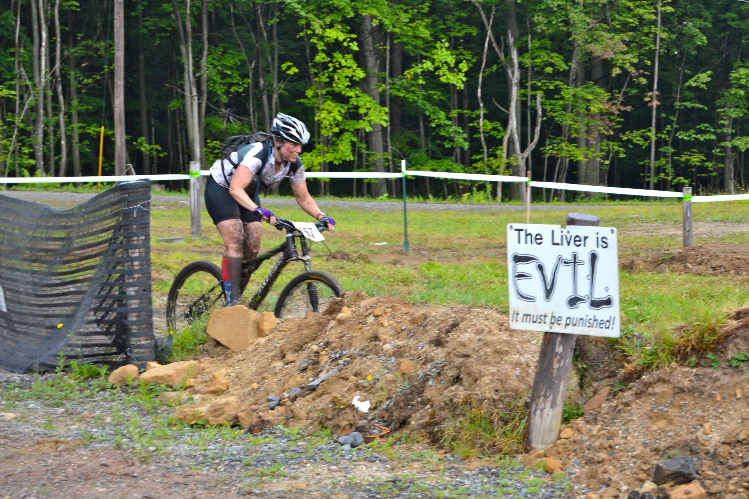 A mountain biker rides through a muddy trail, covered in dirt and sporting a competitive number on their jersey. The background features lush greenery and a playful sign that reads, "The Liver is EVIL! It must be punished!" along with a pile of rocks and dirt on the side of the trail. Big Bear Lake Trail Center mountain bike trail.