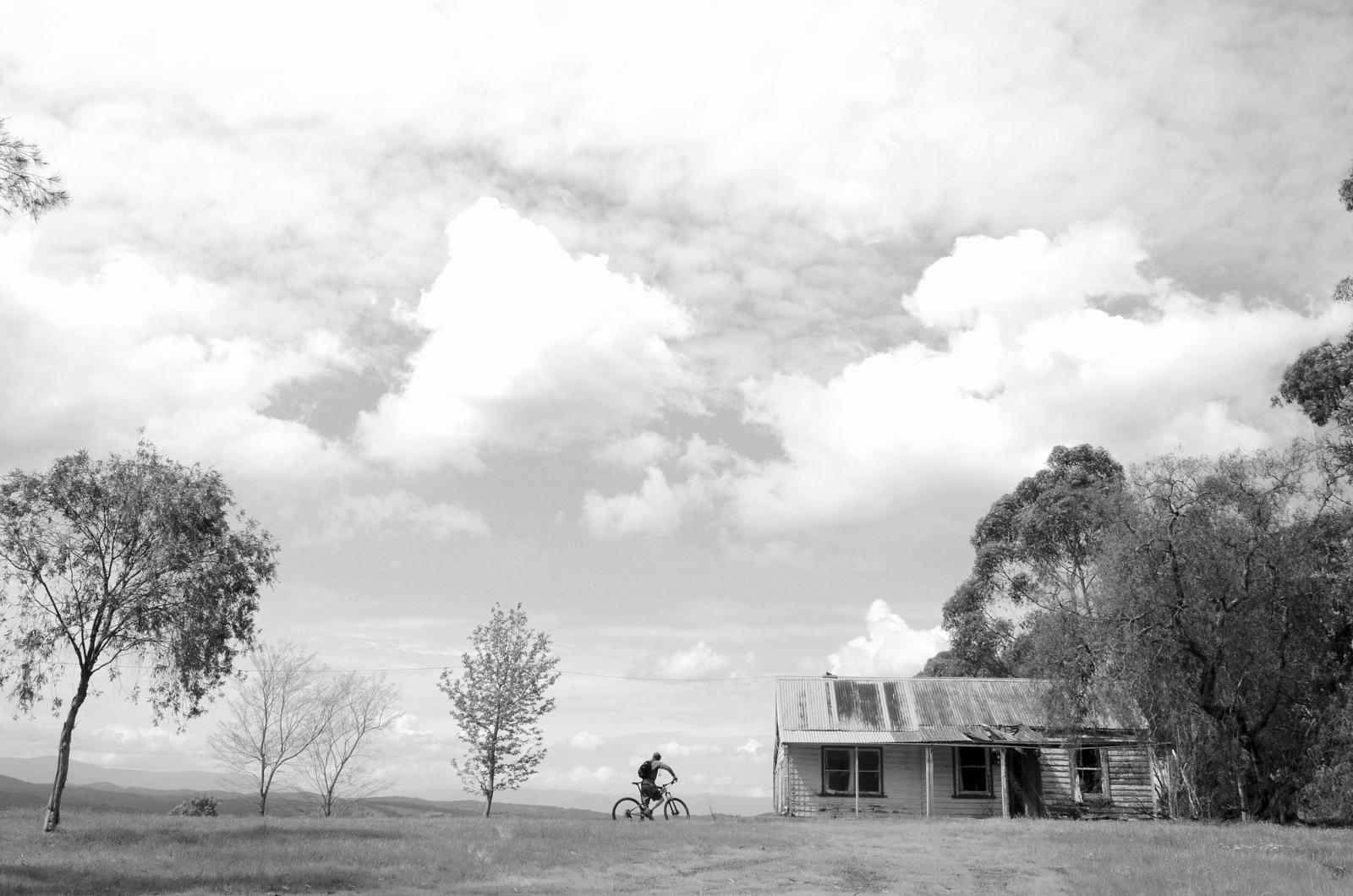 A black and white photograph of a lone cyclist standing beside an old, weathered house, surrounded by sparse trees and rolling hills under a cloudy sky. The scene evokes a sense of solitude and tranquility in a rural landscape. Smiths Gully mountain bike trail.