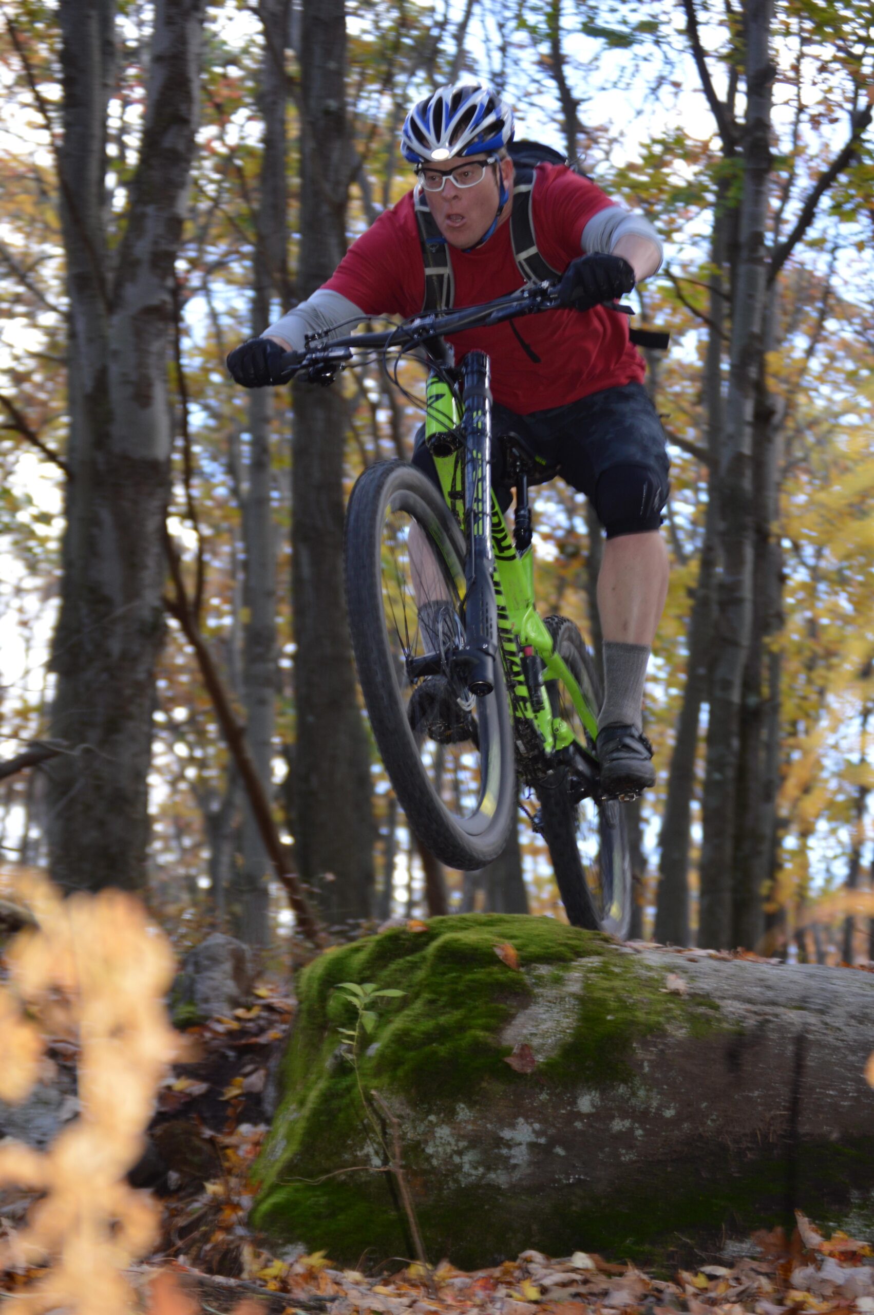 A male mountain biker in a red shirt and helmet jumps off a moss-covered rock while riding through a wooded area with autumn trees in the background. The biker is captured mid-air, showcasing his focused expression and athleticism. Big Bear Lake Trail Center mountain bike trail.