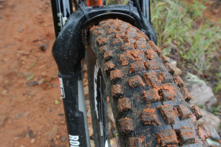 Close-up view of a mountain bike rear tire, showcasing its tread pattern and dirt accumulation. The background features a rugged dirt trail with scattered rocks and green vegetation, indicating an outdoor cycling environment.