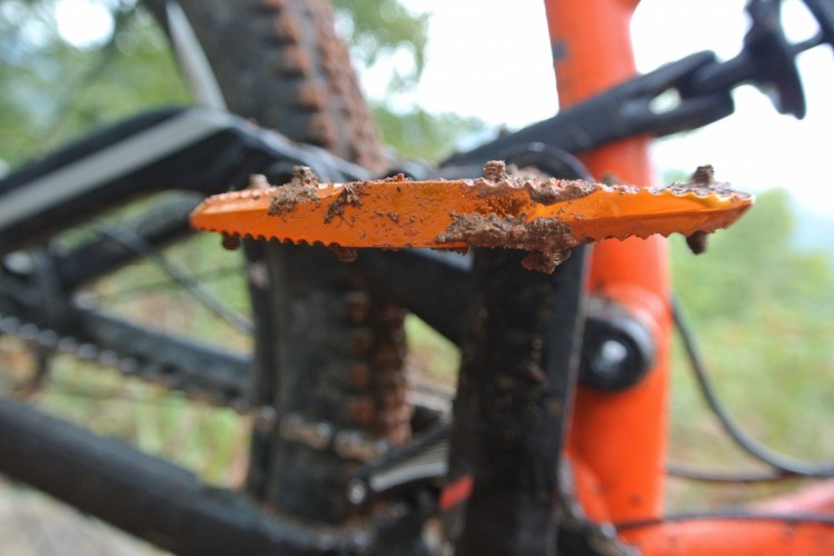Close-up view of an orange bicycle pedal covered in mud, with blurred background showing parts of a bike and greenery.