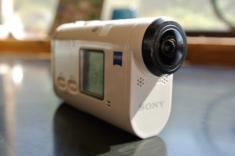 A close-up view of a white Sony camcorder with a Zeiss logo, angled to show its rounded lens and control buttons. The background is softly blurred, indicating indoor lighting.