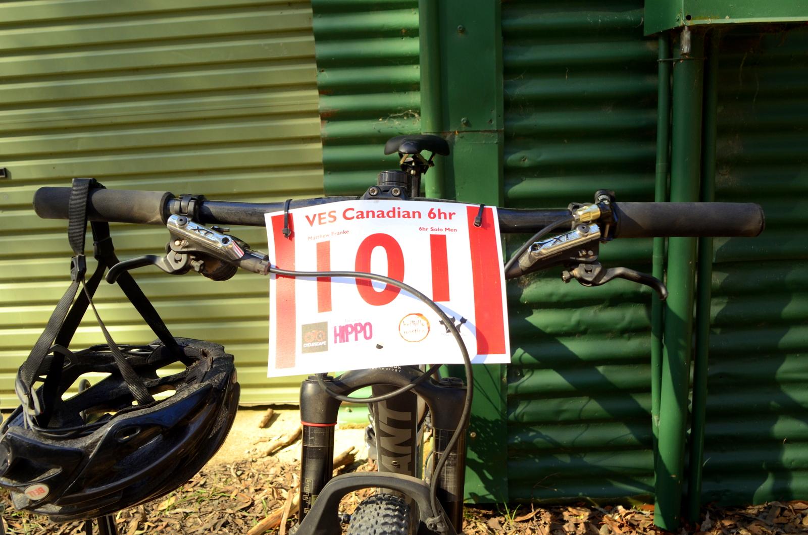 A close-up view of a mountain bike's handlebars, featuring a race number tag labeled "VES Canadian 6hr" with the number "101." The tag also includes the name "Matthew Franke" and details indicating it's for "6hr Solo Men." A black helmet is hanging from the handlebars, and the background shows a corrugated green metal surface. Pax Hill mountain bike trail.