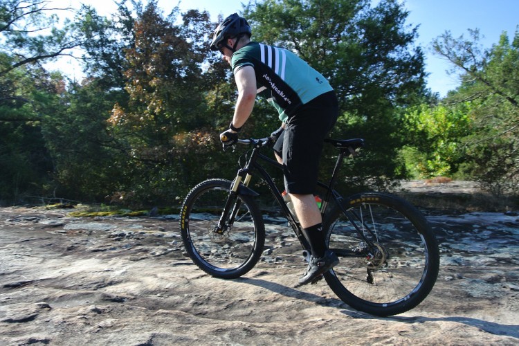 A mountain biker navigating rocky terrain, wearing a helmet and cycling gear, with trees in the background.