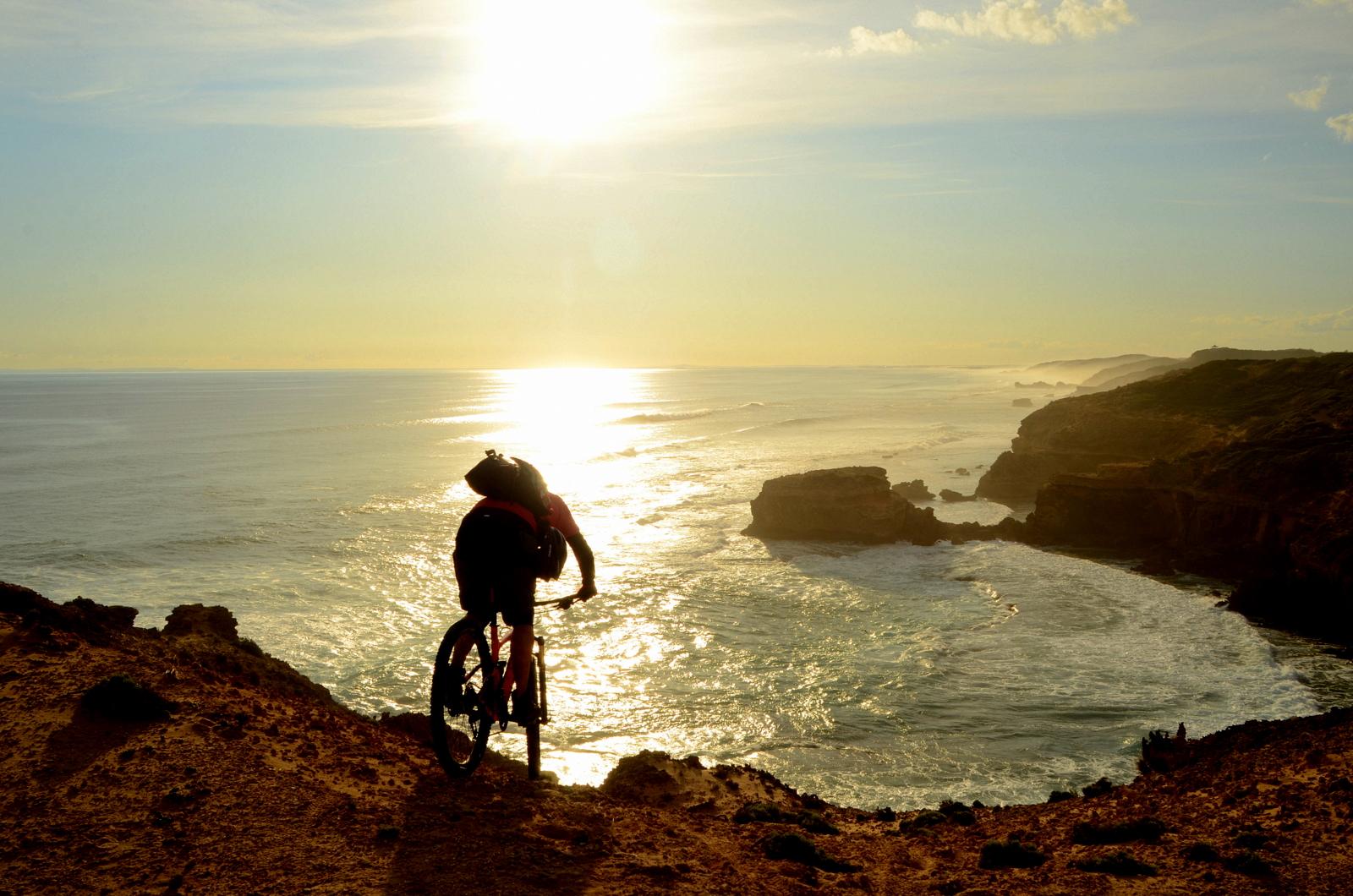 A silhouette of a person riding a mountain bike along a rugged cliff overlooking a calm ocean at sunset. The sun