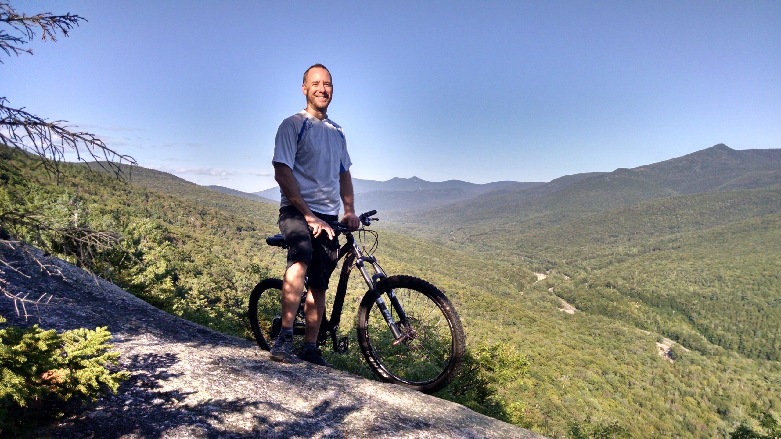 Specialized Stumpjumper FSR Elite: A smiling man stands next to his mountain bike on a rocky outcrop, surrounded by lush green hills and distant mountains under a clear blue sky.