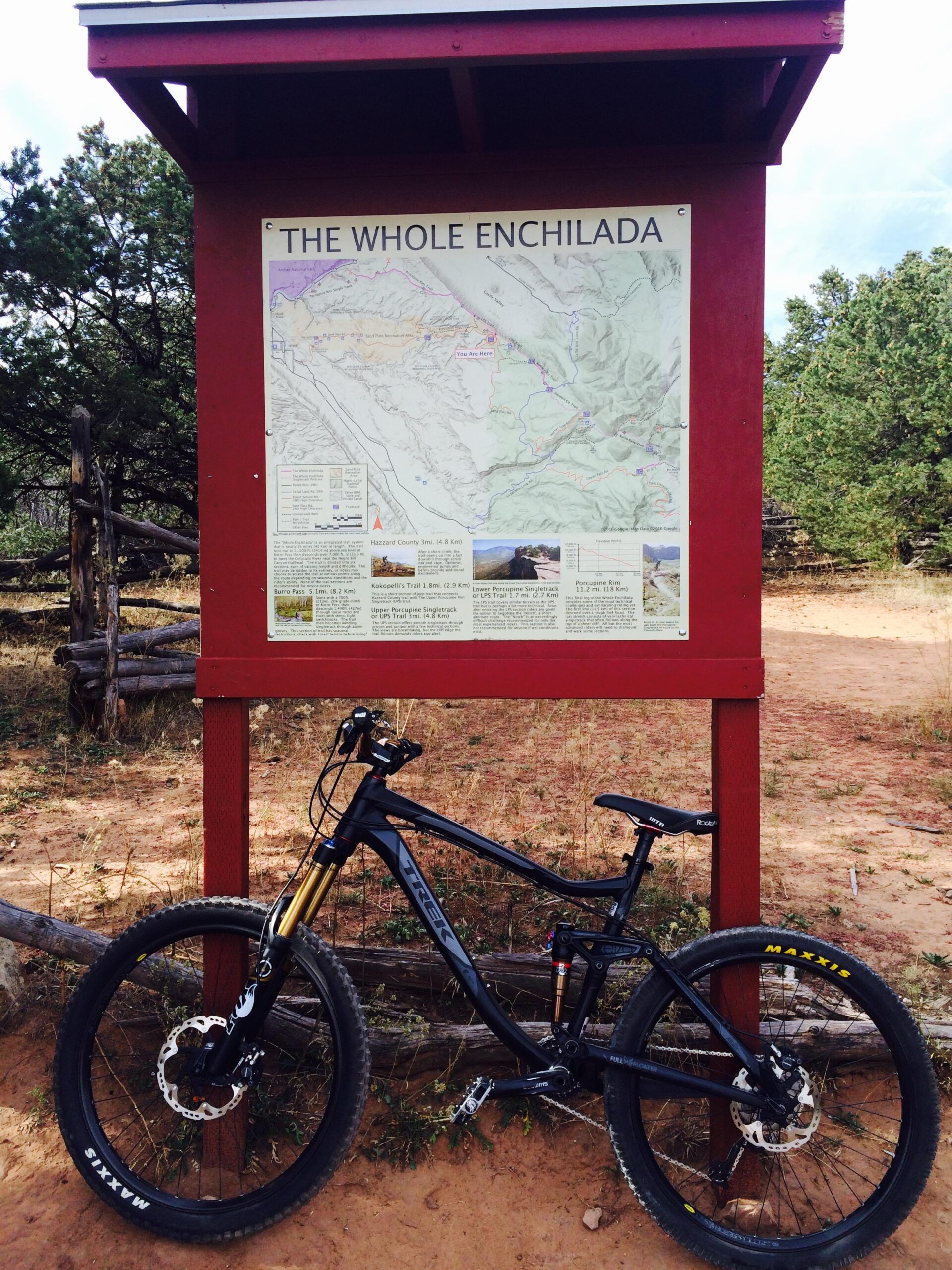 Trek Fuel EX 8: A mountain bike parked next to a trail map sign titled "The Whole Enchilada," set in a natural outdoor environment with trees and dirt ground. The sign features a detailed map and information about various trails in the area.