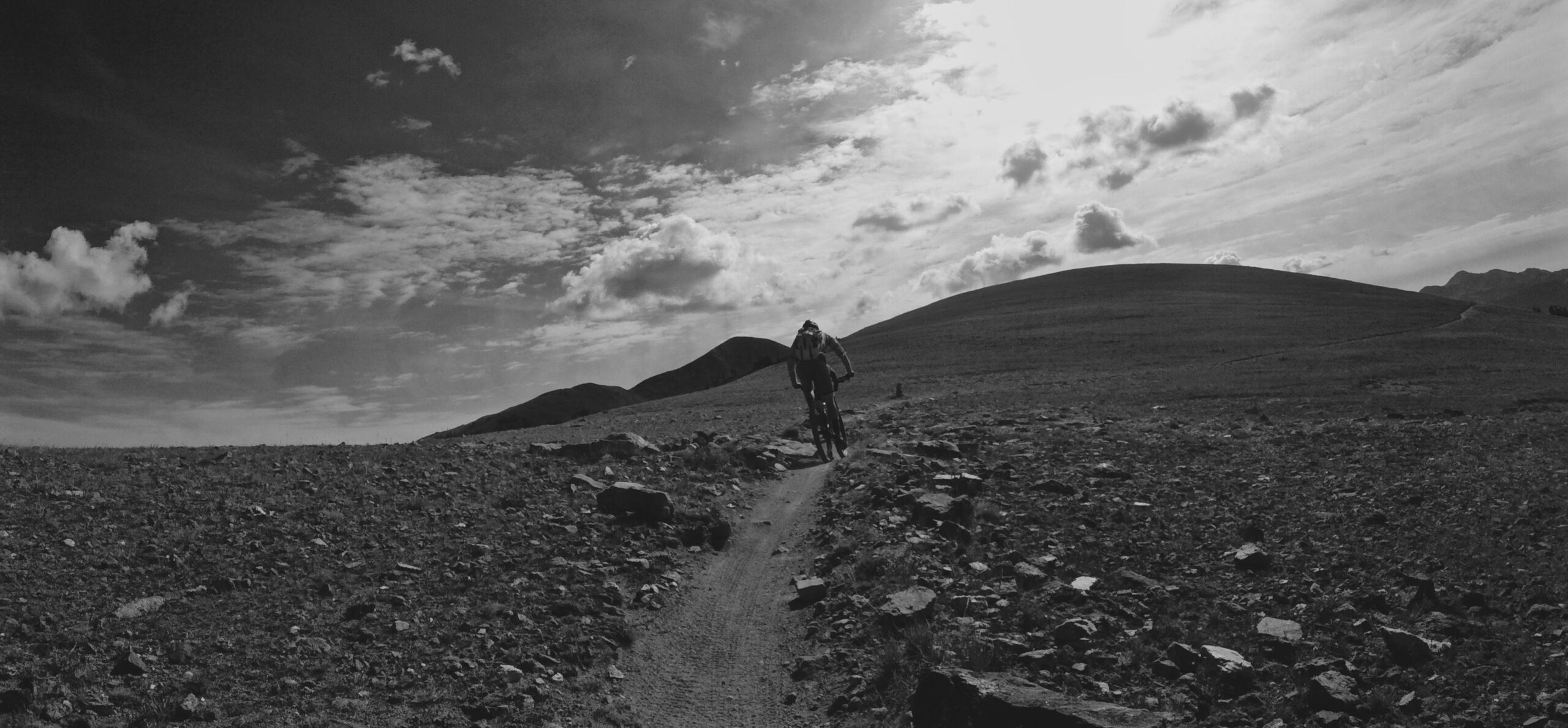 A black and white image of a mountain biker riding along a narrow dirt path surrounded by rocky terrain, with rolling hills in the background and a cloudy sky overhead. The scene conveys a sense of adventure and outdoor exploration. Monarch Crest Trail mountain bike trail.