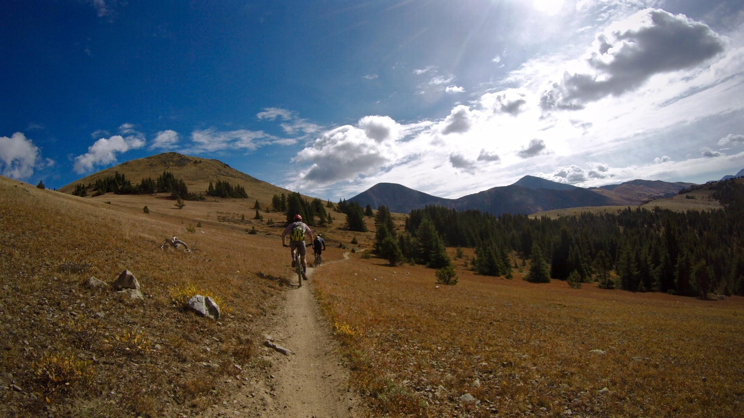 Two mountain bikers ride along a narrow dirt trail through a wide open landscape with golden grass and sparse trees, surrounded by distant mountains under a partly cloudy sky. Monarch Crest Trail mountain bike trail.