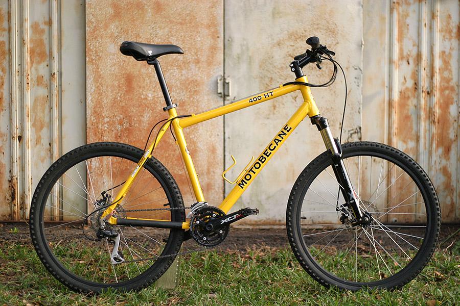 Motobecane 400HT: A yellow Motobecane 400 HT mountain bike is displayed against a rustic metal background, featuring a black seat and handlebars, thick tires, and a visible gear system. The bike stands on grass, highlighting its rugged design suitable for outdoor adventure.
