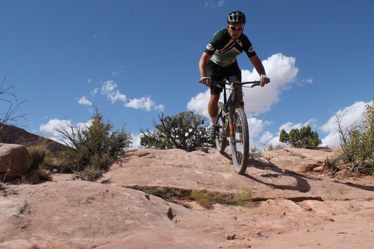 A mountain biker navigating rocky terrain under a blue sky with scattered clouds. The rider is wearing a helmet and sunglasses and is focused on maneuvering the bike over the boulders, surrounded by sparse vegetation.