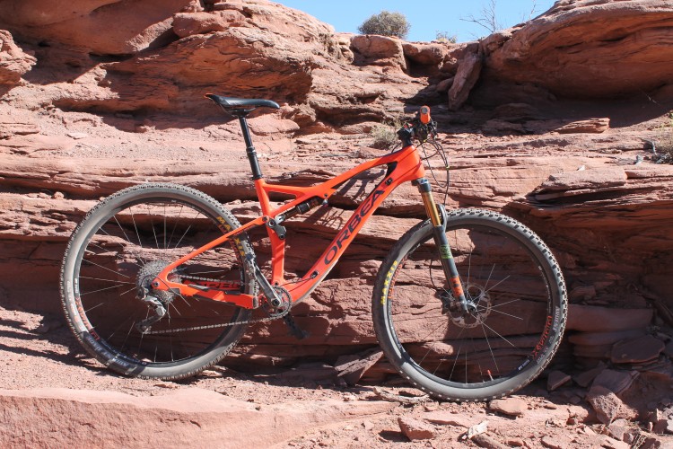 A bright orange mountain bike is positioned against a rugged, red rock formation. The bike features a lightweight frame, front suspension, and knobby tires designed for off-road terrain. The background showcases a clear blue sky and rocky outcrops, emphasizing a natural outdoor setting.