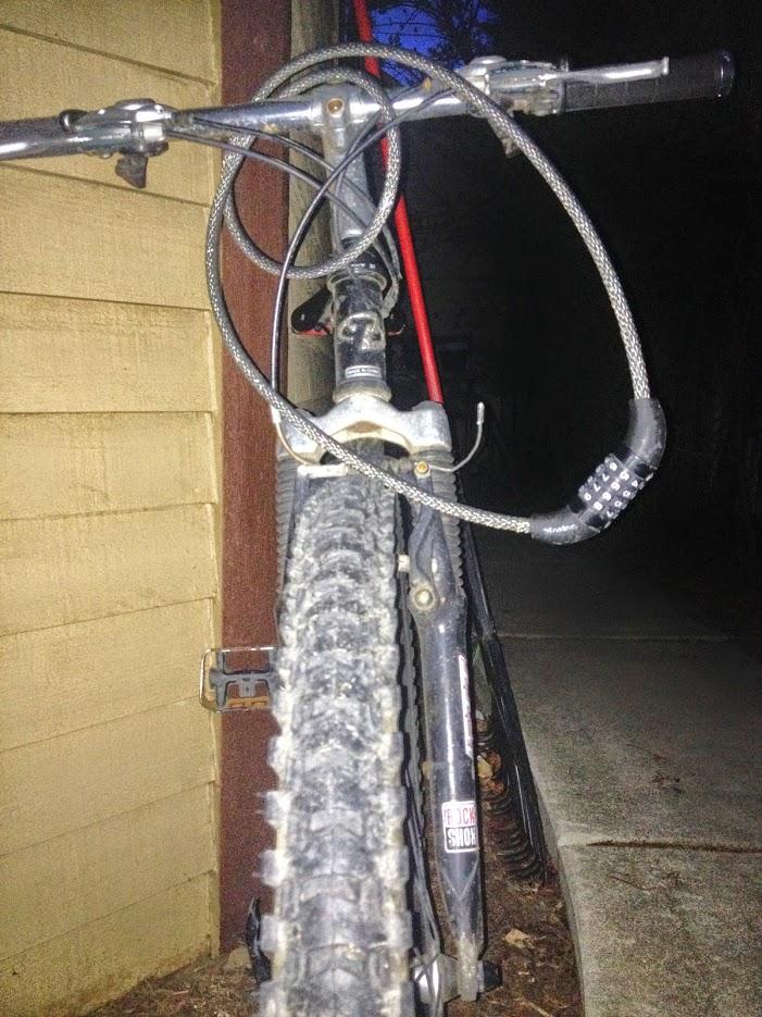 Trek 4500: A close-up view of a mountain bike parked near a building, with its tire in focus. A combination lock is wrapped around the bike's frame, and there is visible dirt on the tires, suggesting recent use. The background is dimly lit, indicating it may be early evening or dusk.