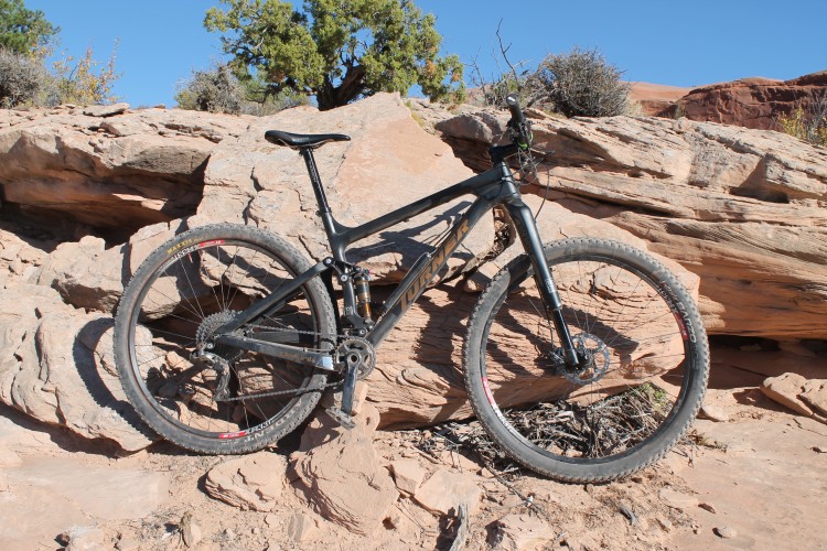 A black mountain bike resting against a large rock formation in a sunny outdoor setting, surrounded by rugged terrain and sparse vegetation. The bike features thick tires and a sturdy frame, indicating it's designed for off-road adventures.