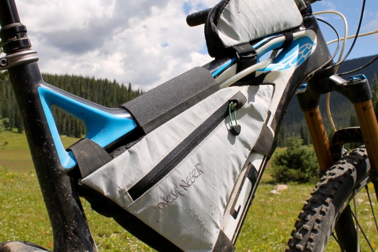 Close-up of a mountain bike featuring a silver and black frame with a blue accent. The bike is equipped with a fitted saddlebag and a top tube bag, designed for carrying gear on outdoor rides. In the background, lush green meadows and a mountainous landscape are visible under a partly cloudy sky.
