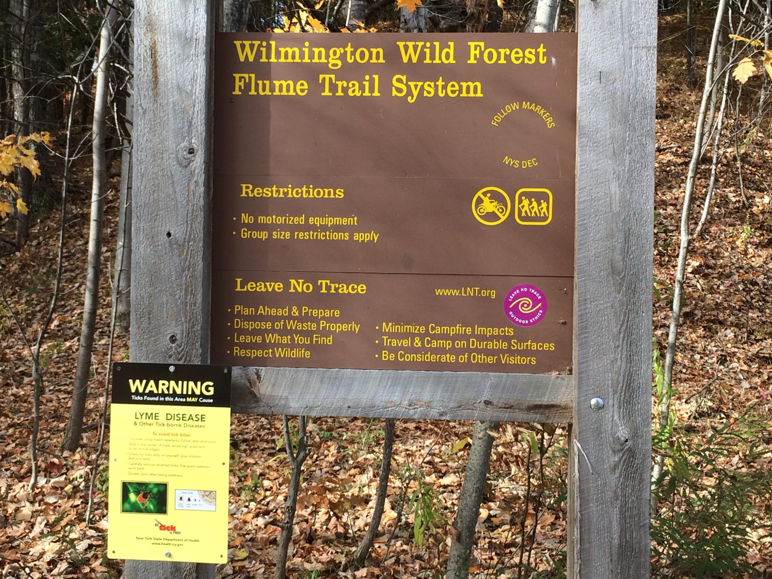Signpost for Wilmington Wild Forest's Flume Trail System, outlining restrictions on motorized equipment and group sizes. It includes "Leave No Trace" principles and a warning about Lyme disease from ticks, emphasizing safety and responsible outdoor behavior. Surrounding environment features autumn foliage. Flume Trails mountain bike trail.