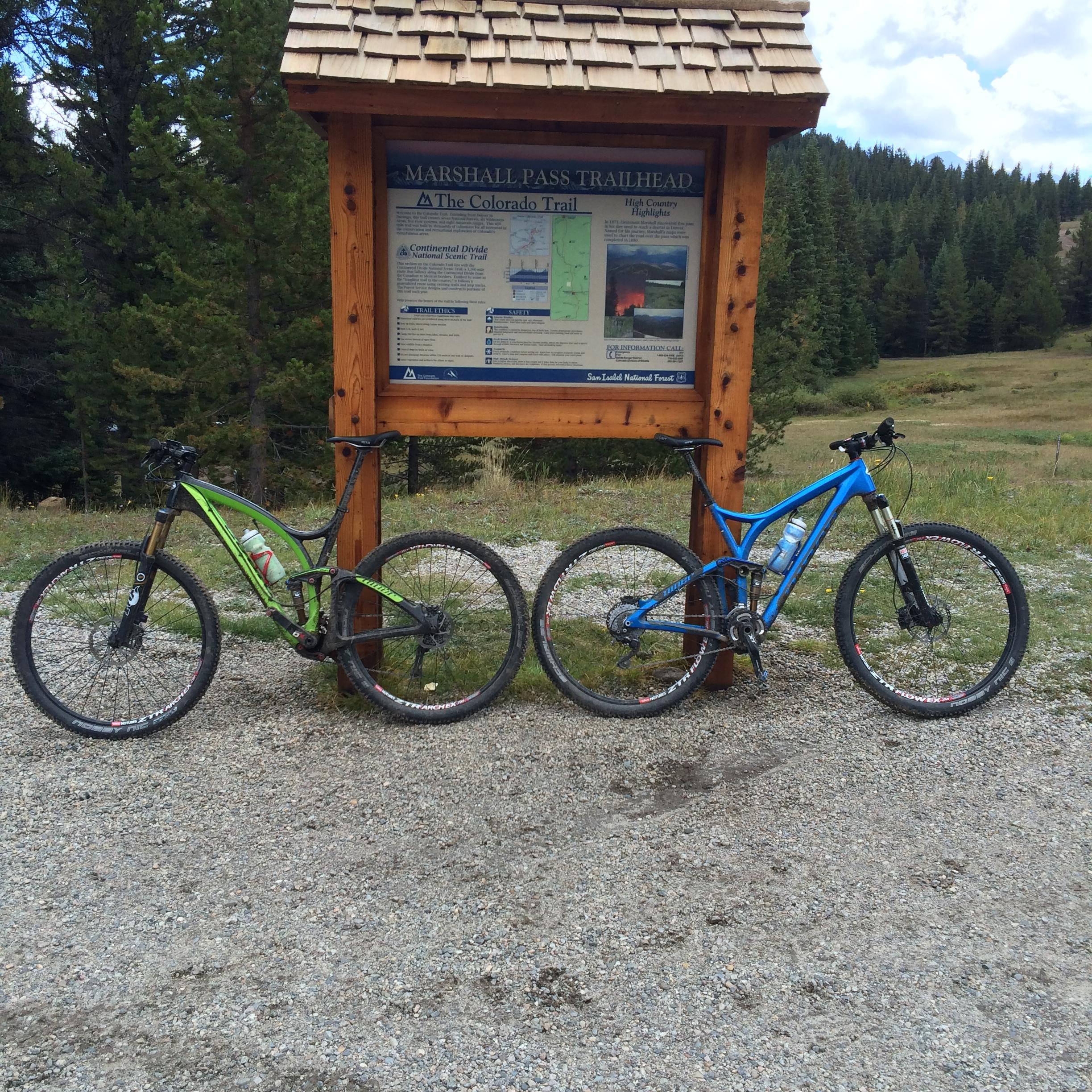 Niner Jet 9 RDO: Two mountain bikes, one green and one blue, are parked next to a wooden sign that reads "Marshall Pass Trailhead" at the Colorado Trail. The background features a grassy area and forested hills under a partly cloudy sky.