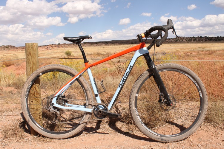 A brightly colored mountain bike with a blue and orange frame is leaning against a wooden post in a rugged outdoor setting. The background features a grassy field and distant rock formations under a partly cloudy sky. The bike has thick tires, showing signs of dirt, suggesting it has been used for off-road riding.