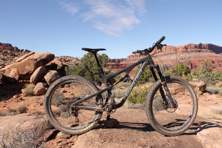 A black mountain bike stands on a rocky surface with a backdrop of red cliffs and blue sky. The bike features a sturdy frame, thick tires, and a visible suspension system, surrounded by sparse desert vegetation.