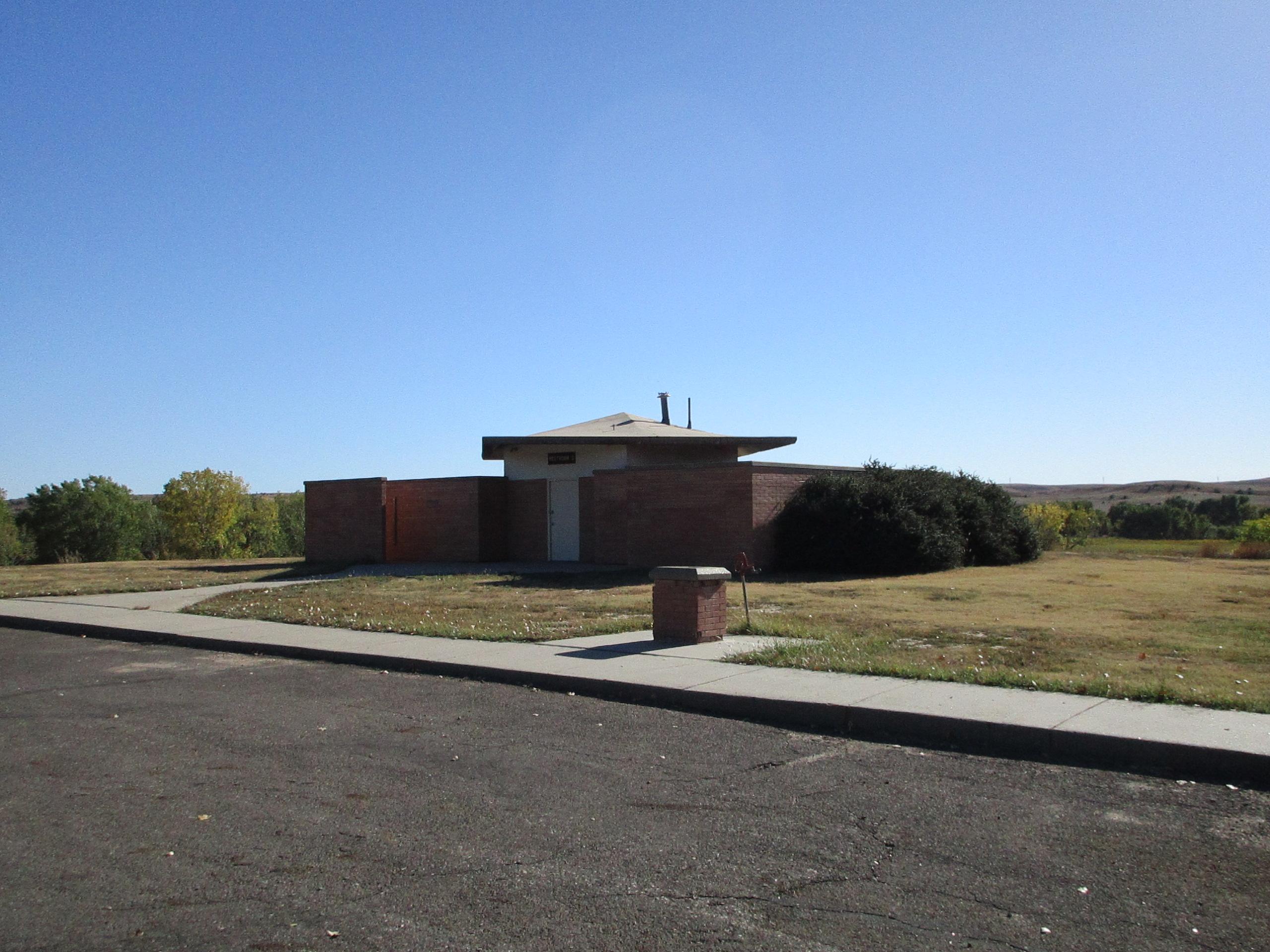 A small, modern brick building with a flat roof, set in a grassy area with sparse trees. The building is positioned near a paved walkway and street, under a clear blue sky. Switchgrass mountain bike trail.