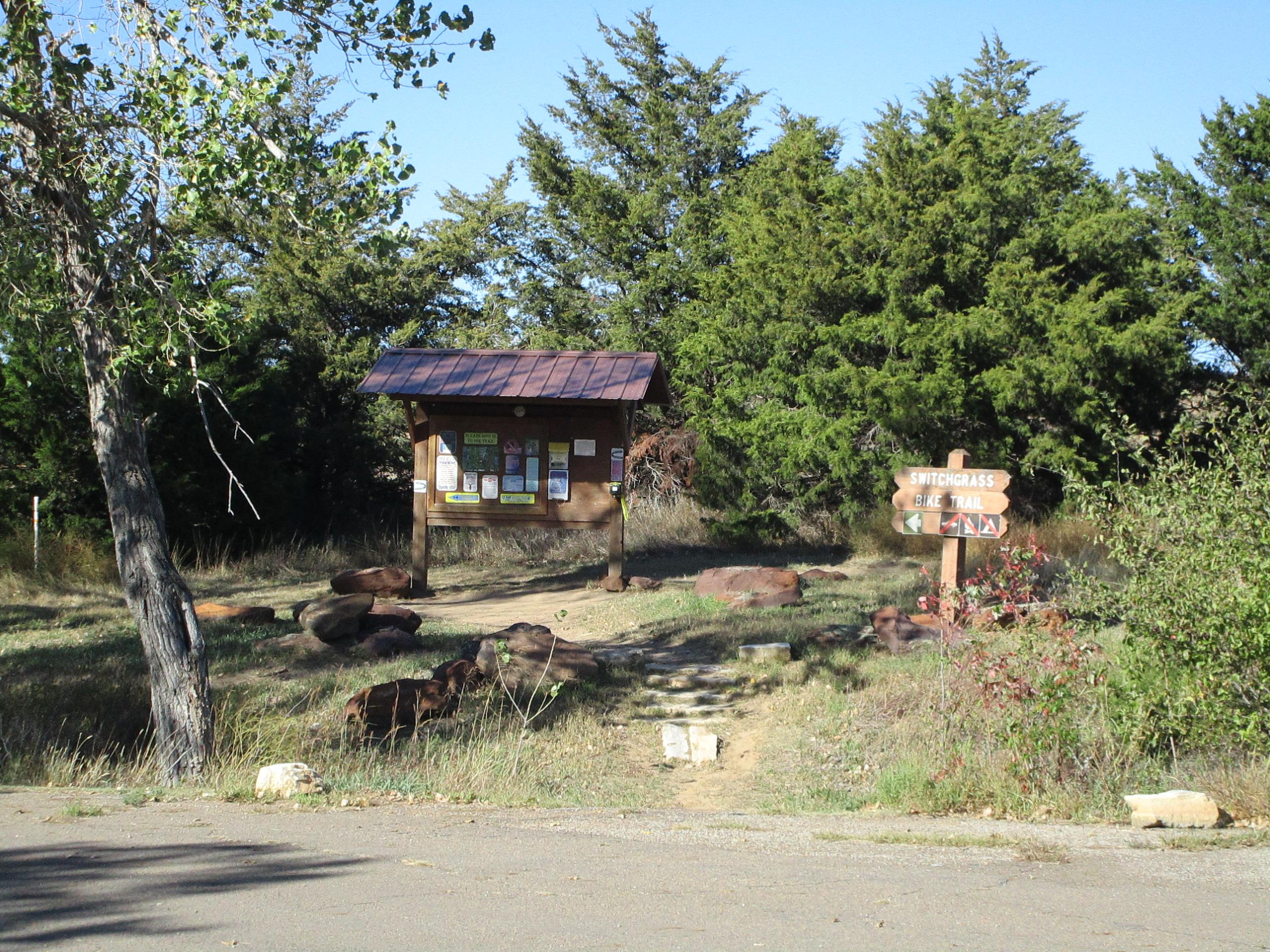 Park trailhead with informational signage and a wooden directional sign. The area is surrounded by greenery, including trees, and features a dirt path leading into the trail. Rocks are scattered around the edge of the path, and the sky is clear. Switchgrass mountain bike trail.