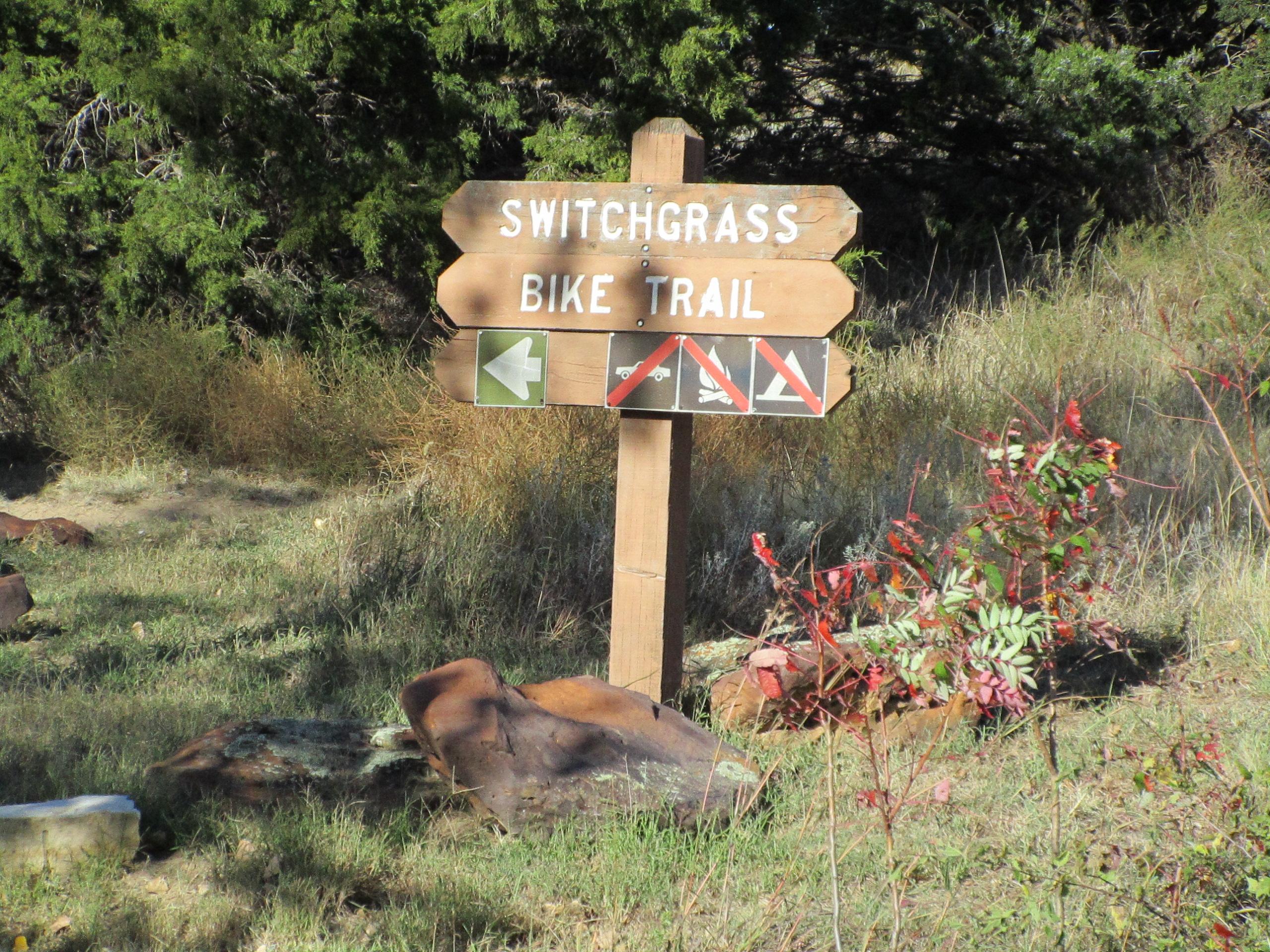 Wooden sign for the Switchgrass Bike Trail, featuring directional arrows and symbols indicating trail use rules, surrounded by grass and small shrubs. Switchgrass mountain bike trail.