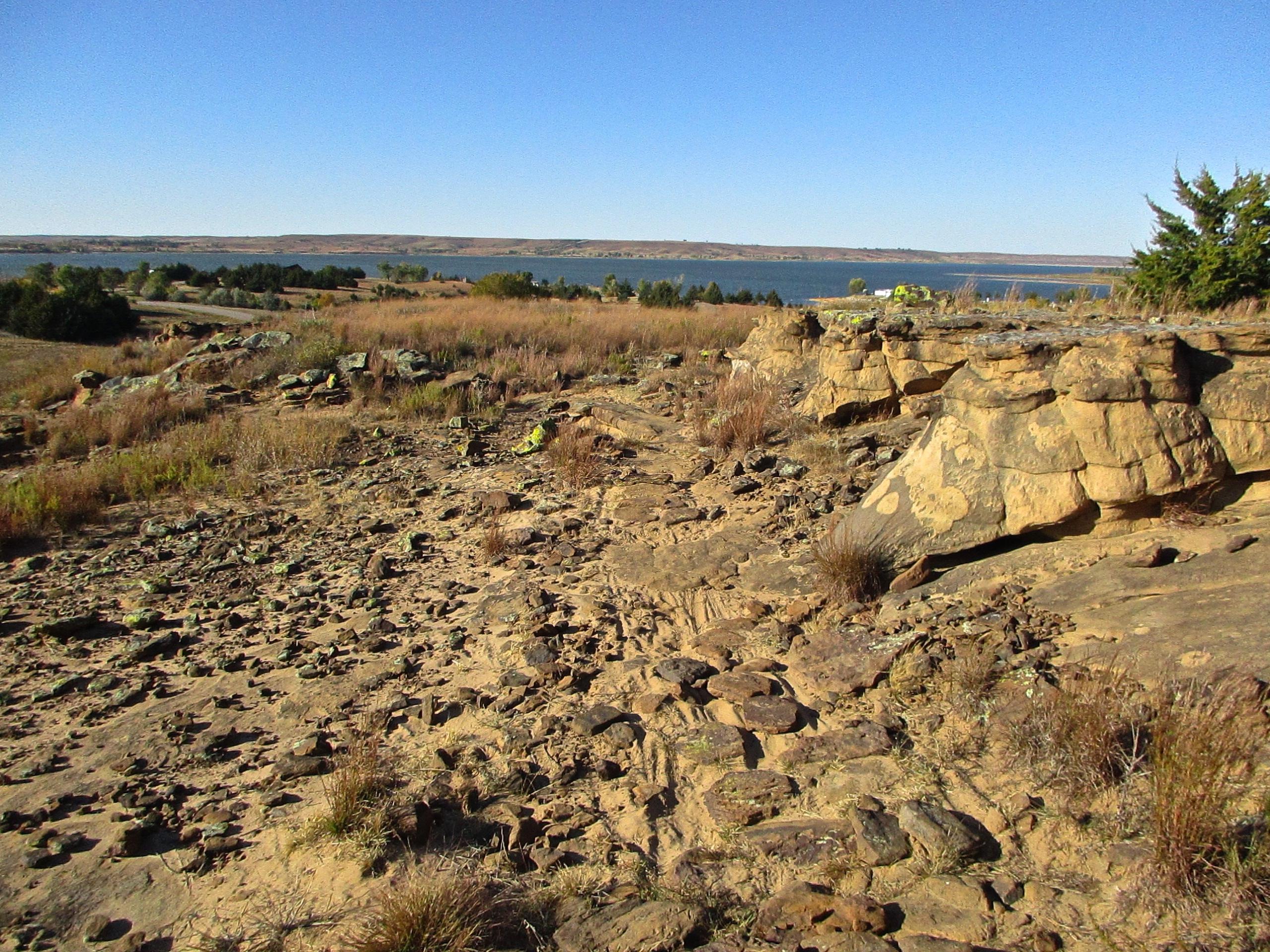 A rocky landscape featuring large, weathered sandstone formations, scattered boulders, and tufts of grass, with a view of a calm lake and distant hills under a clear blue sky. Switchgrass mountain bike trail.