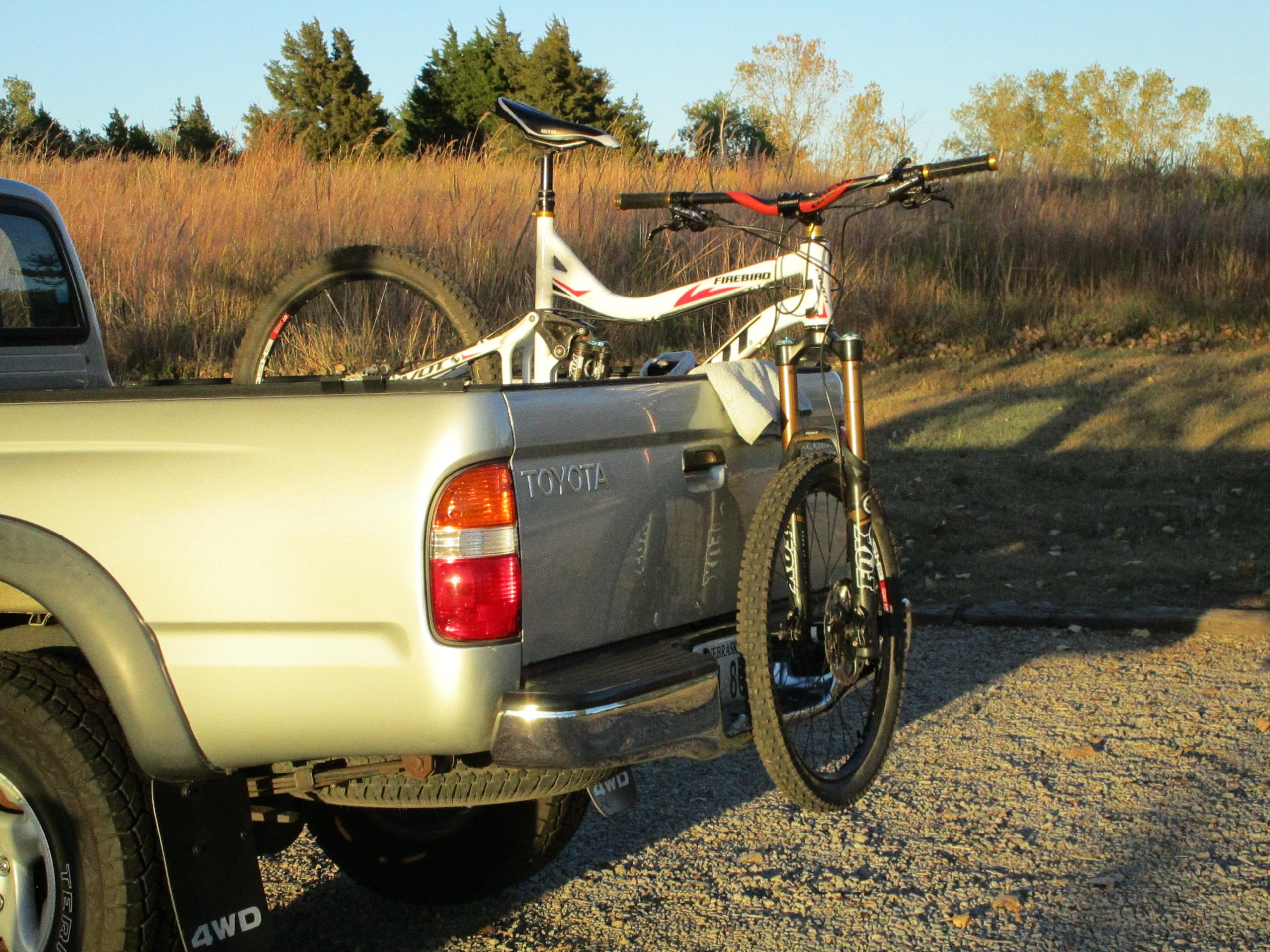 Pivot Firebird 2012: A silver pickup truck is parked on gravel with a mountain bike secured in the bed. The bike has a distinctive design with a white frame and red accents, and its front wheel is resting above the truck