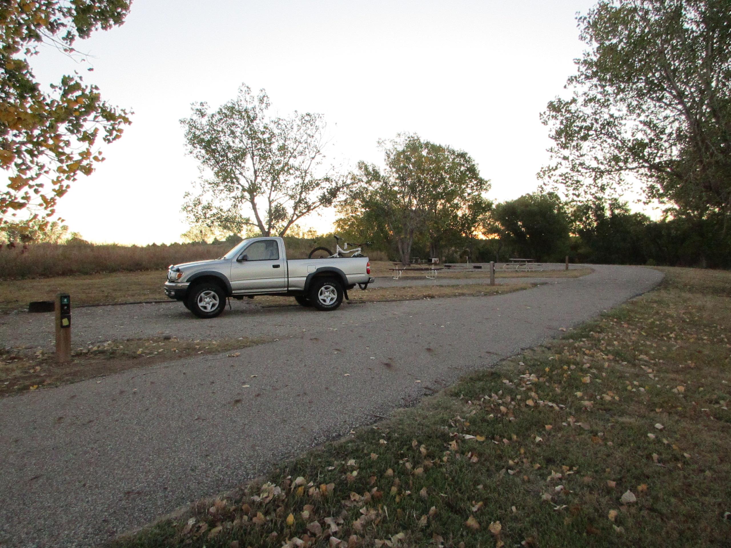 Alt text: A silver truck parked on a winding gravel road in a scenic outdoor area, surrounded by trees and grass with fallen leaves. The background features a picnic area with wooden tables. The sky is transitioning to dusk. Switchgrass mountain bike trail.